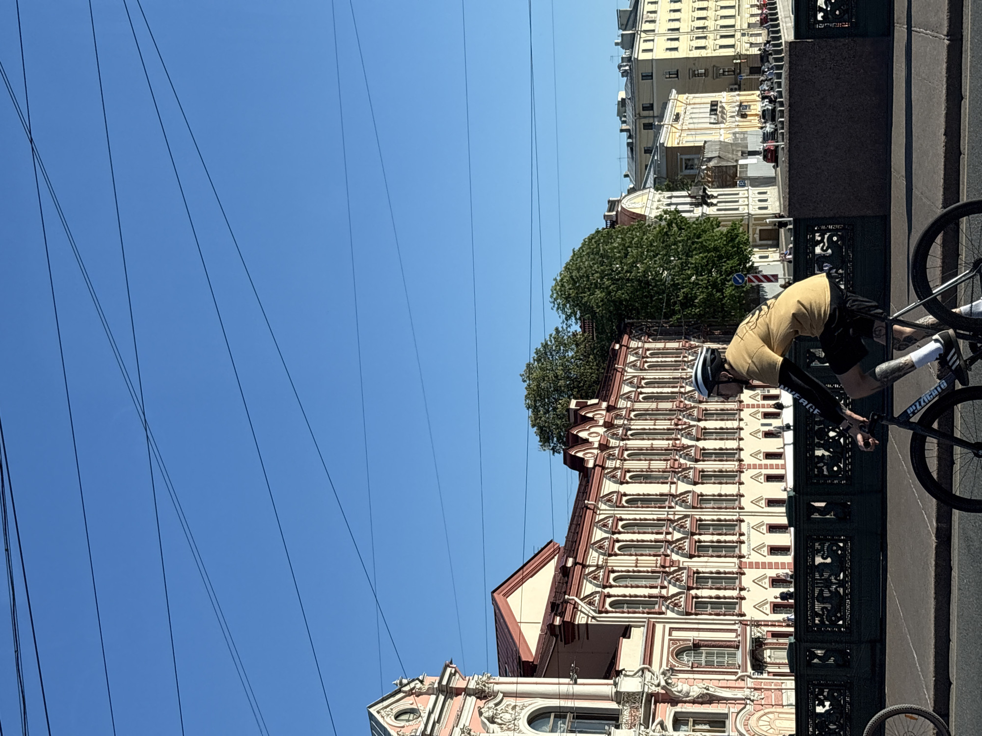 The image depicts a vibrant urban street scene on a clear, sunny day in Saint Petersburg, Russia. The prominent location is the Hermitage Bridge spanning the Winter Canal, with the historic Small Hermitage building visible on the left and other ornate structures lining the embankment.
In the foreground, a male cyclist is riding a black fixed-gear bicycle across the bridge, viewed in profile from the right, moving towards the right. He wears a white helmet, a yellow short-sleeved t-shirt layered over a black long-sleeved shirt, black shorts, white socks with black athletic shoes featuring three white stripes, and has visible tattoos on his lower legs. The bicycle's frame is branded "PIZZICATO." A partial front wheel of another bicycle is visible on the far left edge of the frame.
The bridge itself features an ornate dark-colored cast iron railing with intricate scrollwork and possibly mythological or aquatic creature designs, supported by a granite base. The road surface is asphalt, bordered by a concrete sidewalk with visible seams.
The architectural background is dominated by grand, multi-story historical buildings. On the immediate left stands the Small Hermitage, characterized by its pale pink and cream facade with classical decorative elements, arched windows, and a reddish-brown pitched roof. Behind it, extending across the mid-ground, is a long, cream-colored building featuring numerous regularly spaced arched windows and a matching reddish-brown roof, likely part of the Hermitage complex or an adjacent historical structure. Further in the background to the right, a series of less ornate, multi-story yellow and cream buildings with varied rooflines indicate a distant street where several vehicles are visible. A large, dense green tree is prominently situated between the primary background buildings.
The upper portion of the image is filled with a clear, vibrant blue sky traversed by numerous thin, dark overhead utility lines running both horizontally and diagonally. Small, indistinct figures, presumed to be pedestrians, are visible walking on the bridge and near the background buildings.