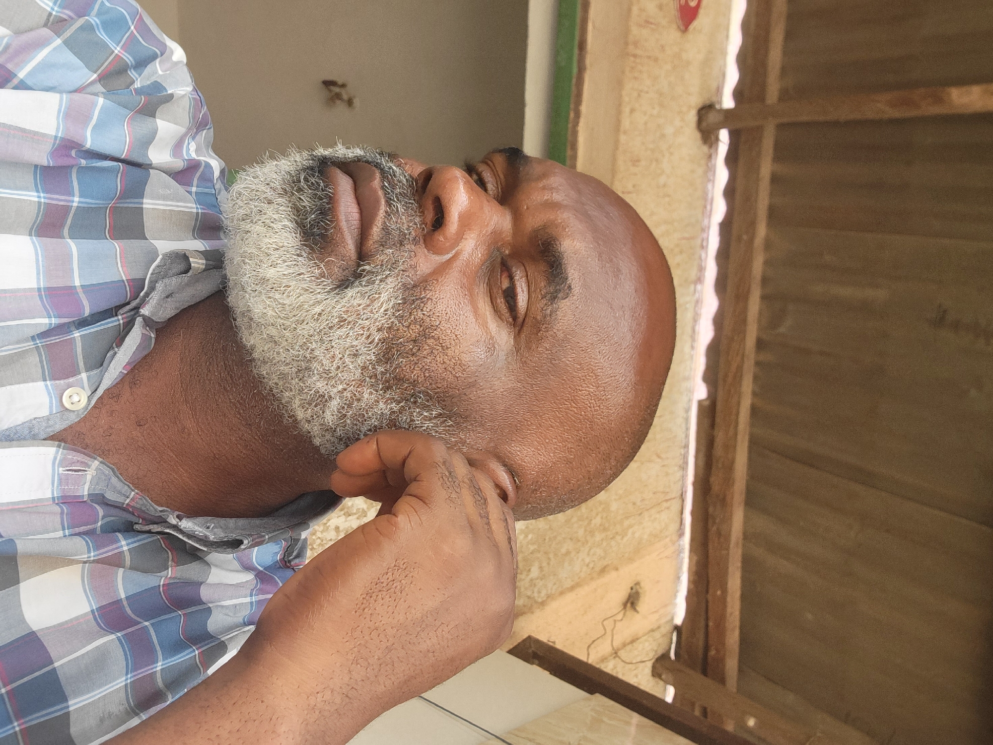 A middle-aged, dark-skinned man is depicted from the chest up, likely indoors, possibly in Maiburiji, Nigeria. He is bald with a full, salt-and-pepper beard. He wears a plaid button-down shirt. His right hand is raised, with fingers near his right ear. The background includes wooden paneling and part of a wall.