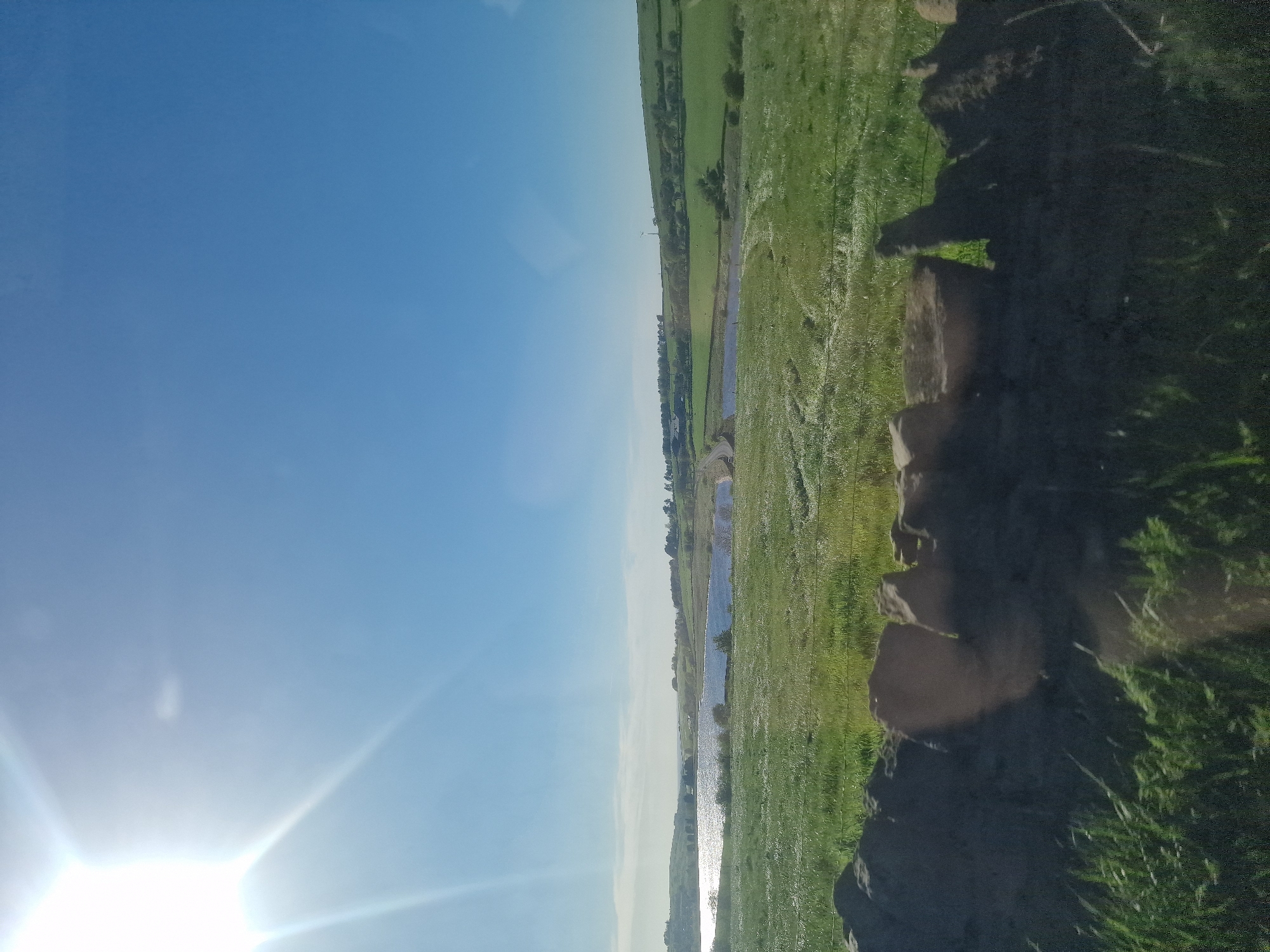 The image displays a wide, sunny landscape under a clear blue sky, viewed from a slightly elevated perspective. The main subject is a pastoral scene featuring vast green fields, a body of water, and a bright sun.
In the upper left portion of the image (when rotated correctly), a brilliant, star-shaped sun flare dominates, indicating a bright, clear day. The sky is a gradient of light to medium blue, with no clouds visible.
The midground features a long, narrow body of water, likely a river or canal, reflecting the sunlight in shimmering streaks. On either side of the water, expansive green fields stretch towards the horizon. The fields are a mix of light and darker greens, suggesting varied vegetation or cultivation. In the far distance, beyond the water and fields, more rolling green hills or fields are visible, extending to a faint horizon line. There might be some distant structures or trees, but they are not clearly discernible due to distance and light conditions.
The foreground of the image consists of a dark, rocky or earthy embankment, which appears to be on the right side of the water body. This embankment is partially covered with patches of grass and shows some irregular rock formations or large clods of earth, creating a textured contrast with the smooth fields and water. The overall impression is one of a tranquil, open-air environment, likely a rural area within Hyndburn, United Kingdom. No human activity or visible text is present.
