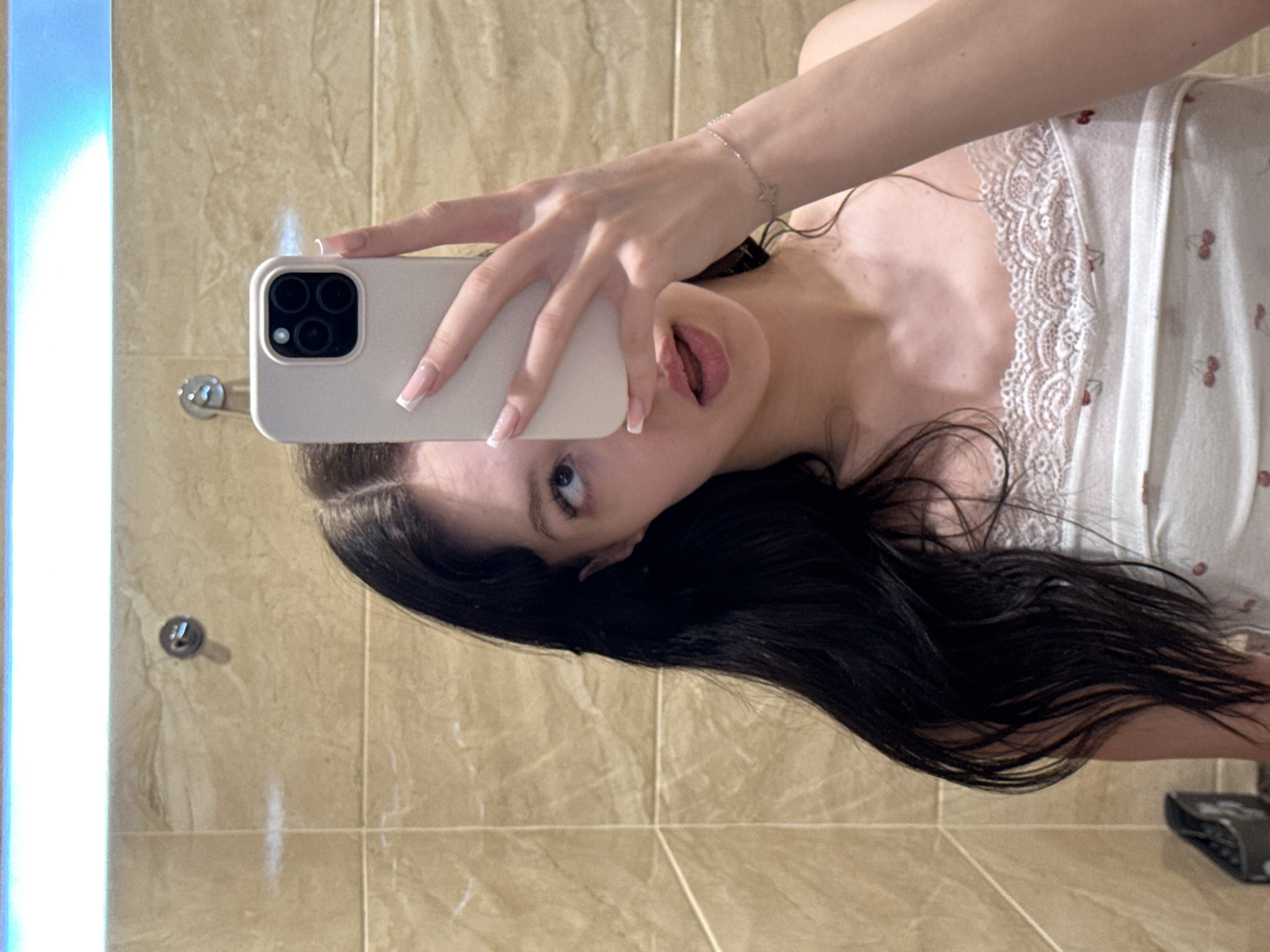 A young woman is taking a mirror selfie in what appears to be a bathroom. She has long, dark hair and is wearing a white tank top with a cherry pattern and lace trim. She is holding a white smartphone in her right hand, positioned to capture the reflection. Her left hand is extended, and she has a bracelet on her left wrist. The background is a tiled wall, and a bright light is visible above the mirror. The overall impression is casual and personal.