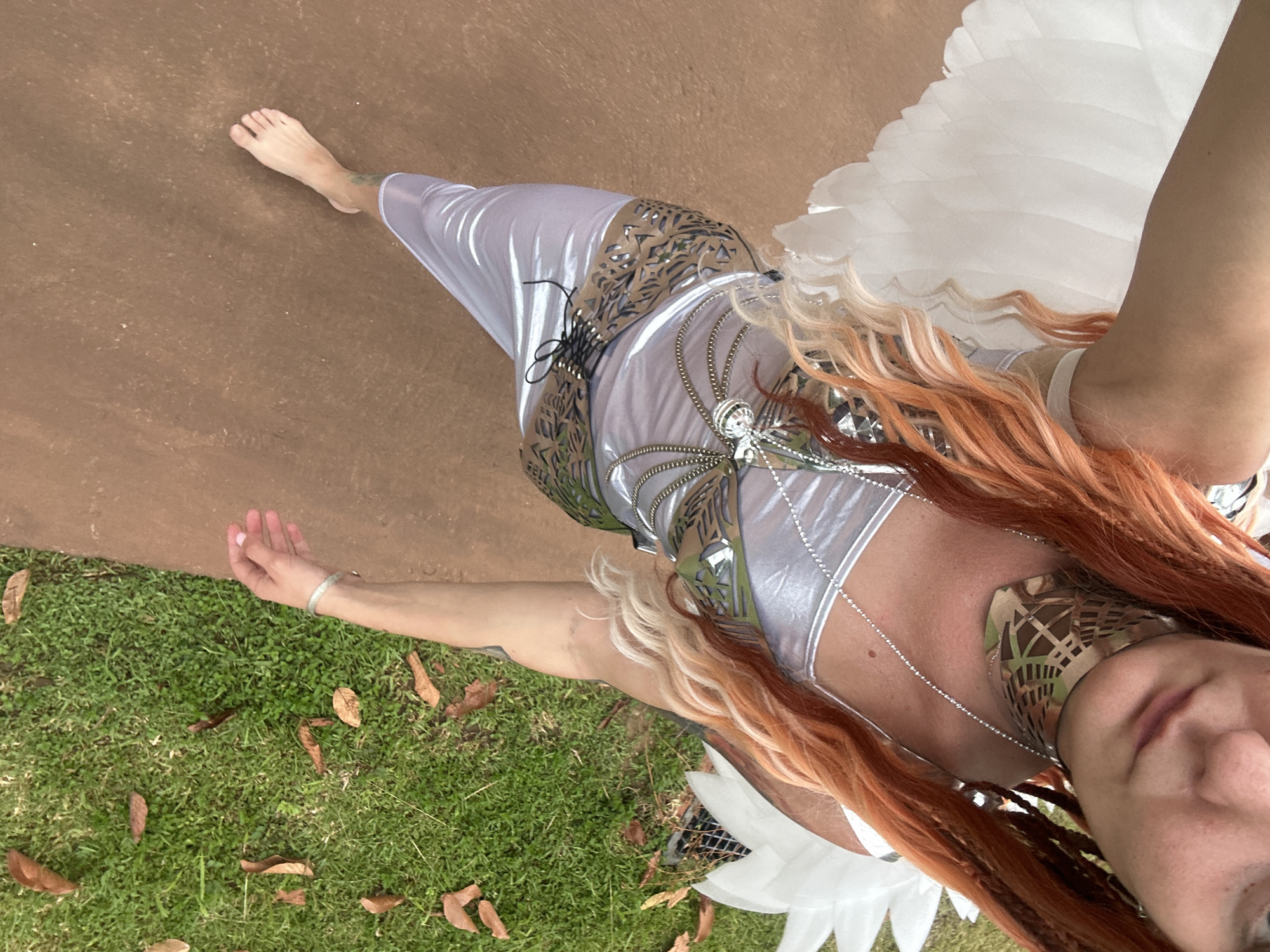 The image shows a person from an overhead perspective, dressed in a futuristic, silvery, form-fitting outfit adorned with a metallic, leaf-like pattern around the waist and chest. The person also has large, white, feathery wings attached to their back. Their hair is long and wavy with strands of blonde and reddish-orange, blending into braids. They are barefoot, with their right foot visible on a reddish-brown surface, possibly a path or court. The lower portion of the image shows a grassy area with fallen brown leaves, suggesting an outdoor setting. The person's left arm is extended outwards, with their hand open, fingers gently splayed. They are wearing a silver bracelet on their left wrist. The lighting and shadows suggest it is daytime. The person's face is partially visible, angled towards the camera, with a neutral or slightly curious expression. The overall impression is one of a fantasy or costume-themed shoot, potentially in a tropical environment given the location context of Ko Samui District, Thailand.