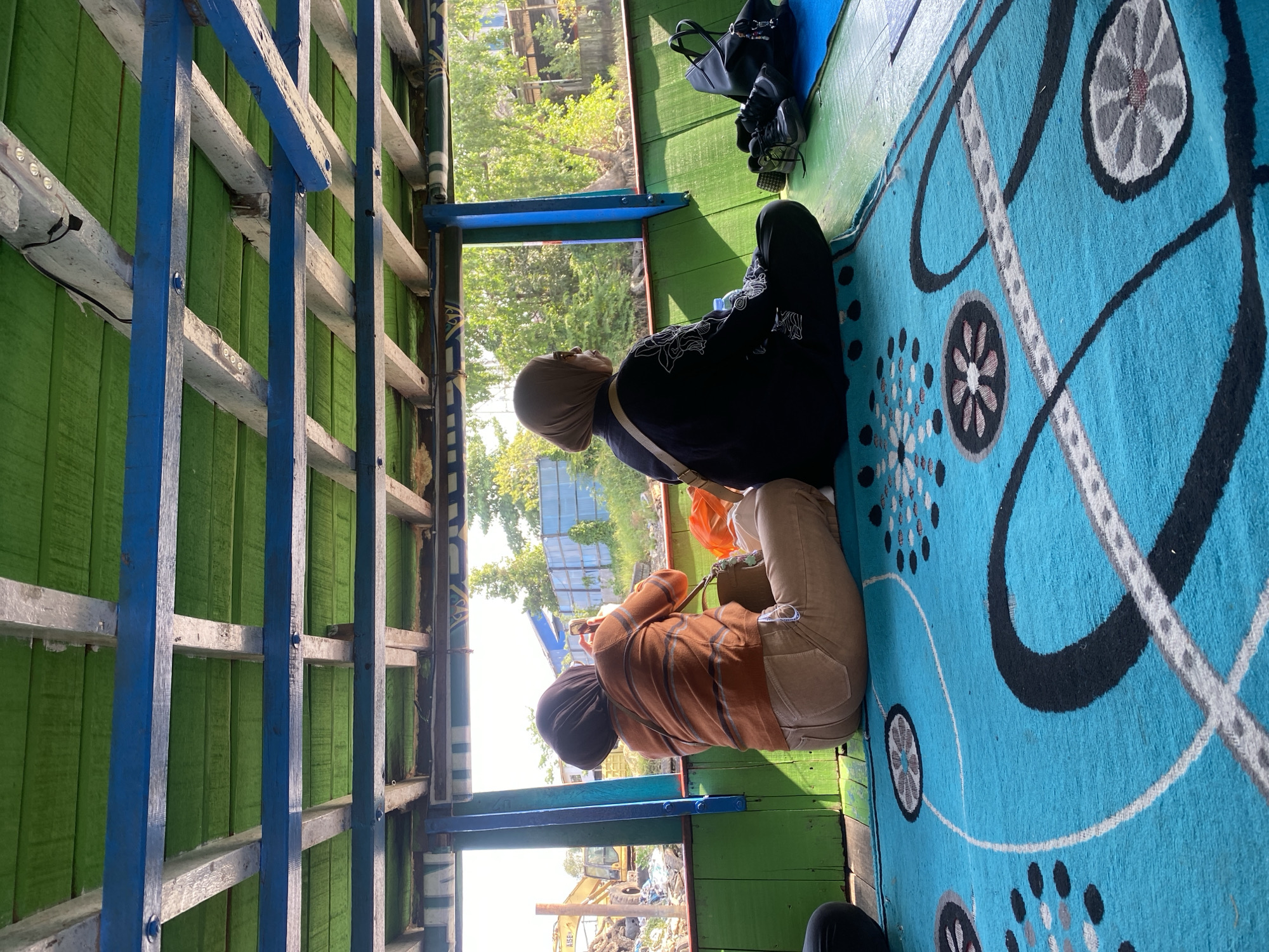 The image depicts two individuals sitting on a blue carpet inside a boat, which is likely in the city of Banjarmasin, Indonesia. The boat's interior features green wooden walls and a ceiling made of green wooden planks supported by blue beams. The person on the left, wearing an orange and brown striped sweater and tan pants, is holding a smartphone, appearing to take a picture or record a video. The person on the right, dressed in a black tunic and a light brown hijab, is looking outwards through a window. A black handbag and a pair of shoes are placed on the floor near them. Outside the boat, through the window, a canal or river is visible, lined with vegetation and some structures, including what appears to be construction debris and a yellow excavator on the left. The scene suggests a mode of transportation typical of Banjarmasin, a city known for its extensive network of rivers and canals.