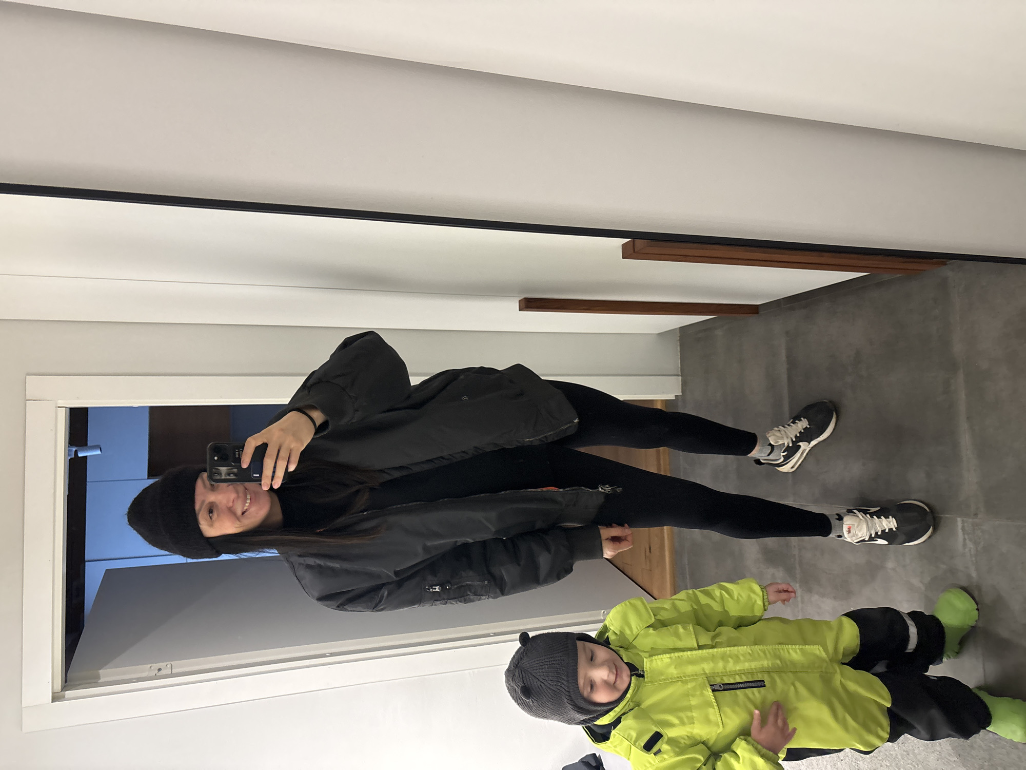 An unspecified indoor location in Chaikovsky, Russia, features a full-length mirror capturing two individuals. On the right, a female adult, estimated mid-20s to mid-30s, is reflected, holding a black smartphone to capture the image. She wears a black knit beanie, a black bomber jacket with an orange lining visible near a zippered sleeve pocket, a black high-neck top, black leggings, and black and white athletic shoes (Nike brand visible). Her expression is a smile. To her left, a young child, approximately 2-4 years old, stands looking downwards and slightly left, with a subtle smile. The child wears a grey knit hat with two small ear-like protrusions, a bright neon-yellow full-body snowsuit with black lower leg sections and white reflective stripes, and bright neon-green rubber boots. The floor consists of dark grey square tiles. Walls are light-colored, and a white door frame is visible in the background, leading to a darker interior space with blue-tinted reflections and a ceiling light fixture. The mirror has a thin dark frame and two vertical wooden elements reflected on its surface.