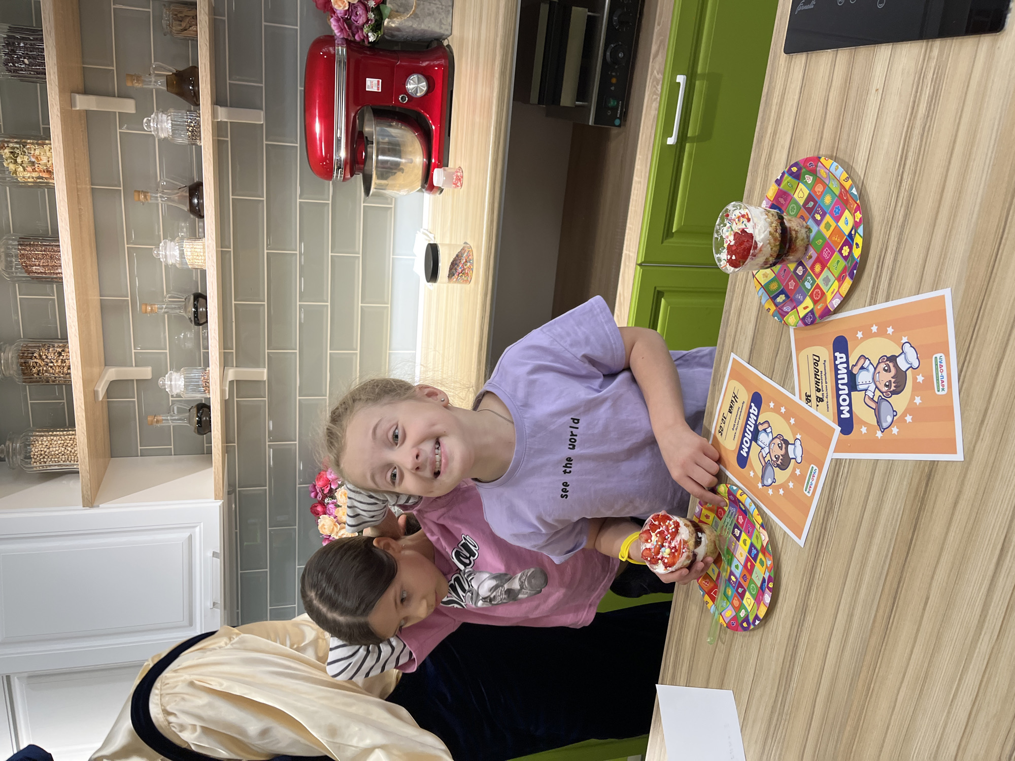 Two girls are in a kitchen, smiling and holding desserts and certificates. The girl in the purple t-shirt is in the foreground, smiling at the camera, while the other girl in the pink t-shirt is slightly behind her. Both girls are holding desserts in cups and colorful plates, and there are two certificates lying on the counter with the word "ДИПЛОМ" (Diploma) written on them, which appear to be awards for completing a cooking course. In the background, there is a kitchen setup with shelves containing jars of food, a red stand mixer, and green cabinets. The room appears to be well-lit.