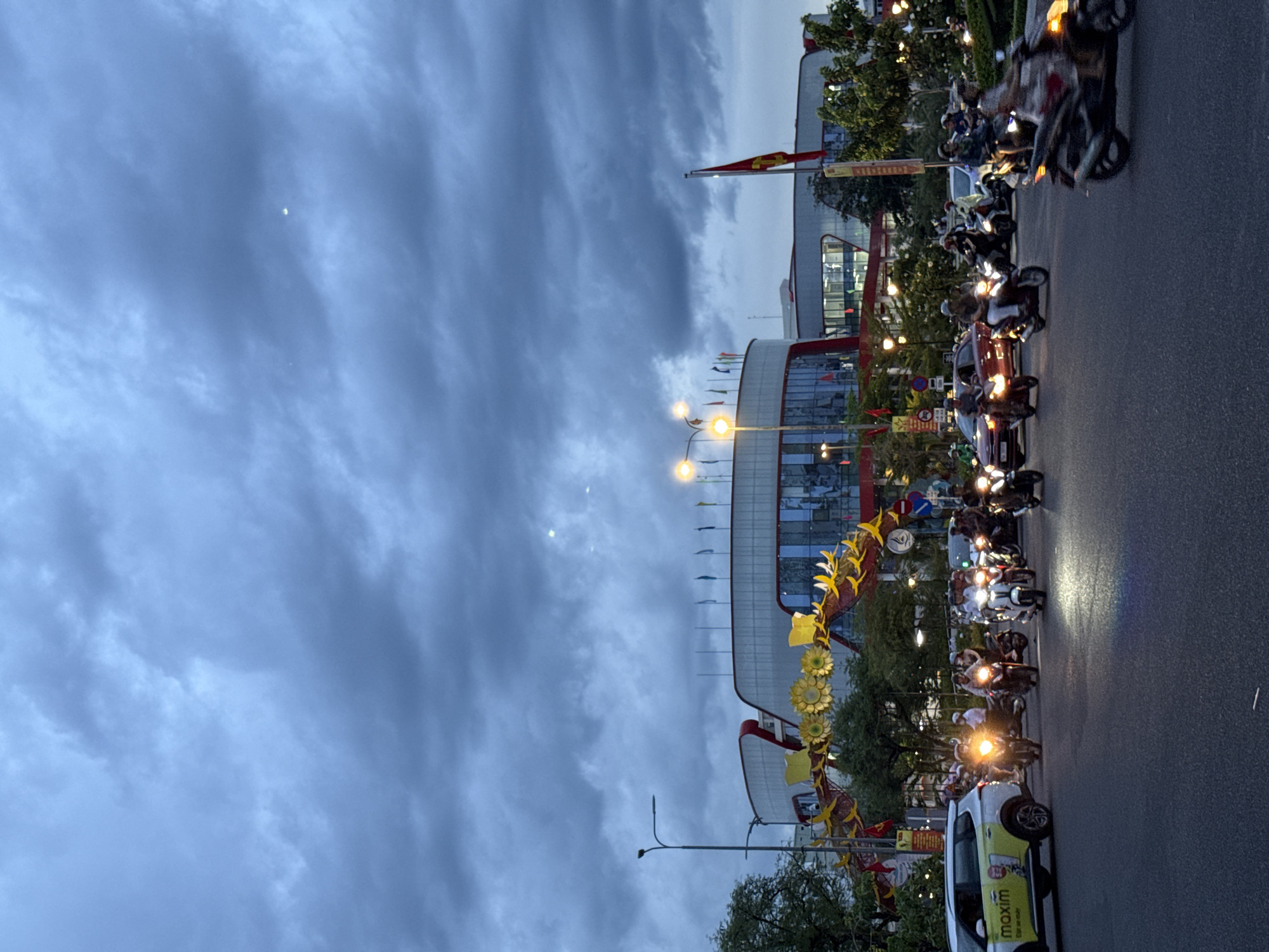 The image depicts a street scene in Nha Trang, Vietnam, during what appears to be early evening or twilight. The sky is overcast with dark, dramatic clouds. In the background, a modern building with a rounded facade, featuring red accents and large glass windows, is visible. Flags are displayed along the roofline of the building, and a red flag with a yellow emblem flies from a flagpole to the right. A large, illuminated arch decorated with yellow sunflowers and dragon motifs spans across the street in front of the building. The foreground shows a busy intersection with numerous motorcycles and a few cars navigating the road. Many of the motorcycles have their headlights on, illuminating the asphalt. People are visible riding the motorcycles, some wearing helmets. A white SUV with yellow branding that reads "maxim" is parked on the left side of the road. The overall atmosphere suggests a lively urban environment transitioning into night.