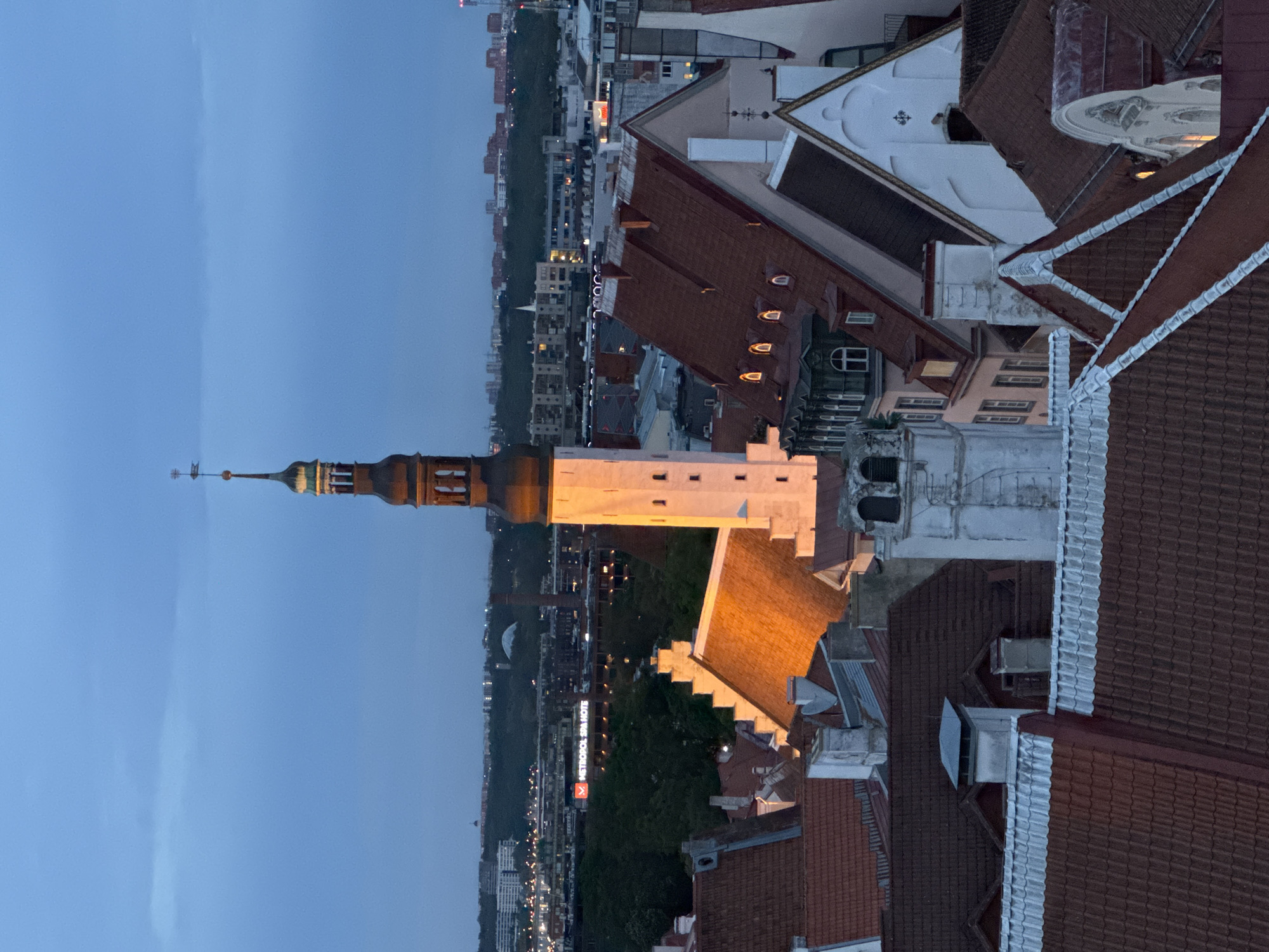 This is an aerial, medium shot of the Tallinn Old Town in Estonia at dusk. The image is dominated by the terracotta tiled roofs of historic buildings and a tall, illuminated church tower with a spire. The sky is a deep blue, indicating twilight. The tower, which appears to be St. Olaf's Church, is lit from within, casting a warm glow on the surrounding buildings. In the background, the modern skyline of Tallinn is visible, with some buildings and lights illuminated. The scene is serene and captures the architectural beauty of the city transitioning from day to night. There are no people or animals visible in the image.