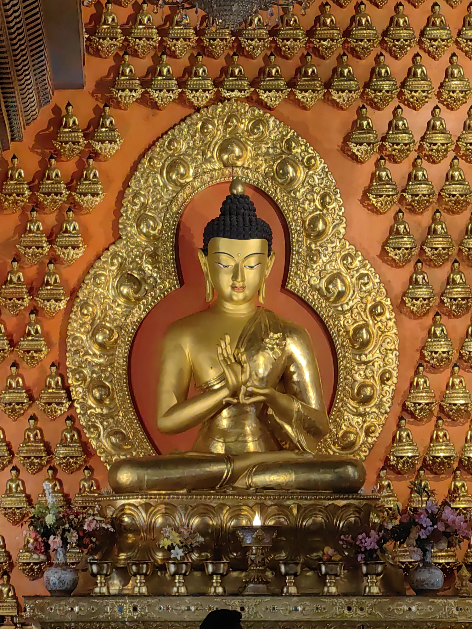 The image depicts a large, golden statue of Buddha seated in a meditative pose, adorned with intricate gold ornamentation. The statue is centrally positioned within a larger, ornate gold halo against an orange backdrop. The background is densely populated with numerous smaller golden Buddha figurines, arranged in a grid-like pattern. In front of the main statue, a tiered altar displays offerings including flowers in a vase, small golden chalices, and a lit oil lamp. The overall scene suggests a religious or spiritual setting, likely a temple or shrine, evoking a sense of reverence and tranquility. Based on the provided metadata, this image was taken in Parsa, Nepal.