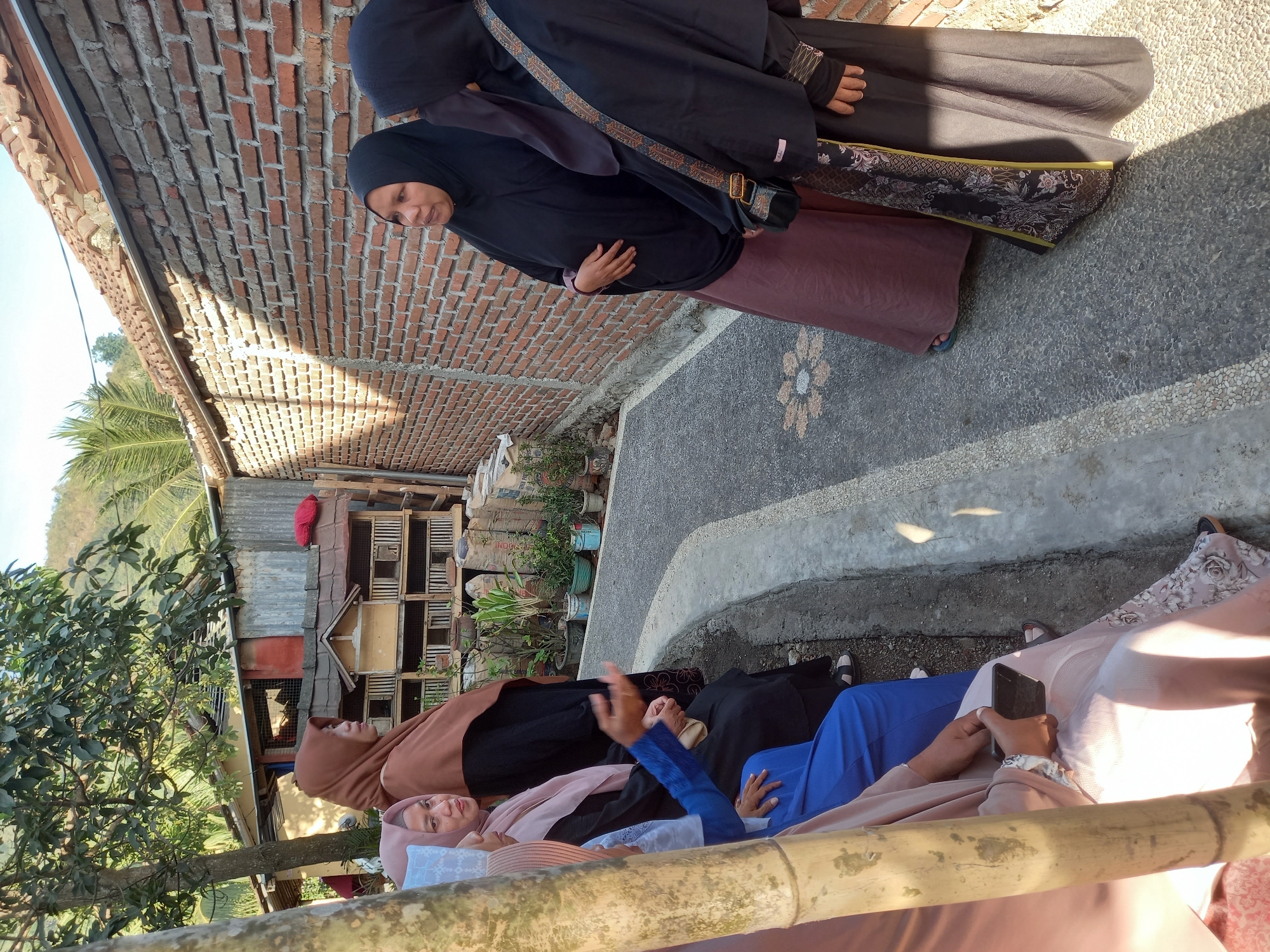 In a residential alleyway in Tempos, Indonesia, a group of women dressed in hijabs are gathered. Two women on the left are seated, one in a light pink hijab and the other in a darker blue abaya. The woman in the pink hijab holds a black smartphone. Two other women stand on the right, facing each other. One woman, in a black hijab and a mauve skirt, gestures with her hand as she speaks. The other woman, in a black hijab and a patterned black and gold skirt, listens intently. Behind them, a brick wall runs along the alley, with potted plants and sacks of materials visible near its base. A small, multi-compartment wooden structure, possibly for animals, is also situated nearby, shaded by trees. The overall scene suggests a casual outdoor gathering in a semi-rural setting.