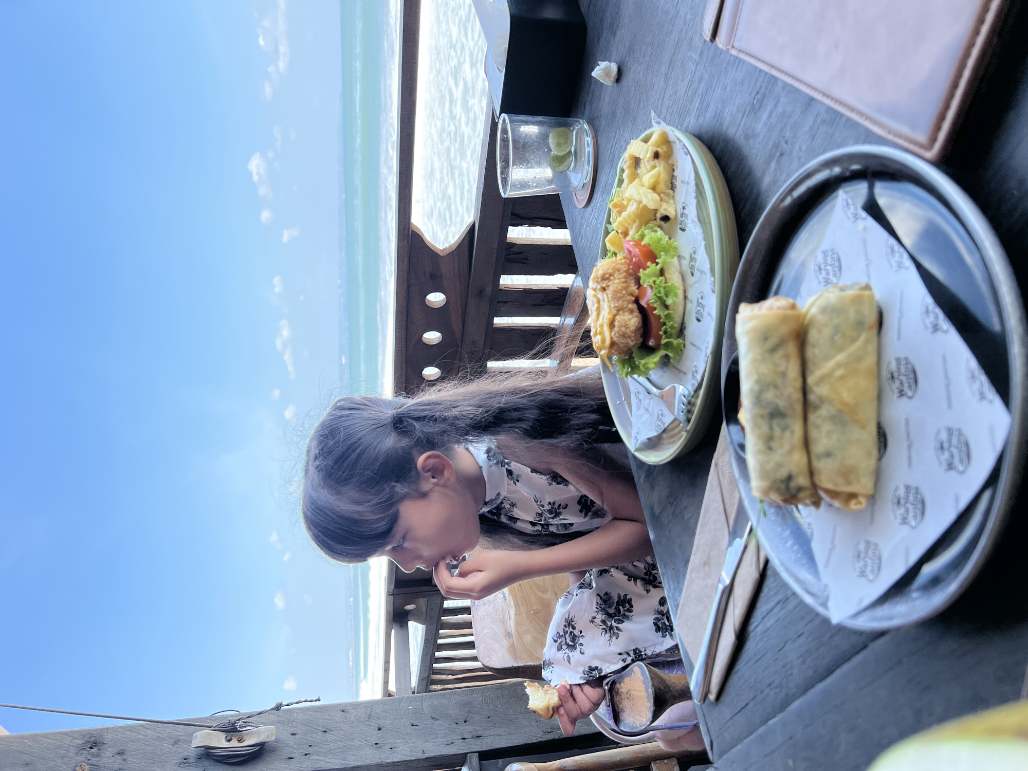 A young girl with long, dark hair pulled into a ponytail sits at a wooden table outdoors, facing right and eating a piece of food. She is wearing a white dress with a black floral pattern. In front of her, on the table, are two plates of food: one with two spring rolls and another with a burger and fries. In the background, the ocean is visible with waves crashing on the shore under a bright blue sky. The setting appears to be a restaurant or cafe in North Kuta, Indonesia, suggested by the beachside location and Indonesian architecture of the railing.