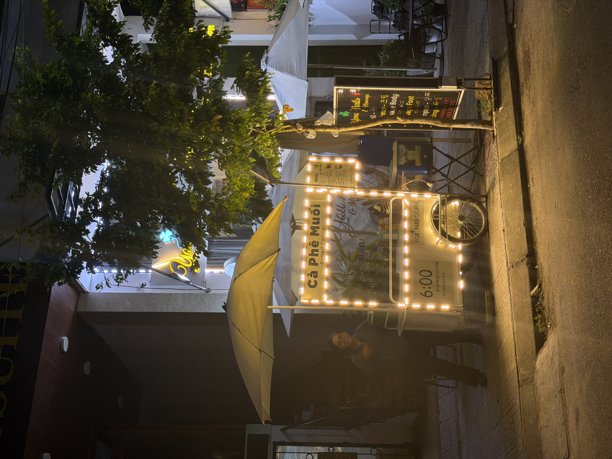 The image is taken in Đà Nẵng, Vietnam, at night. A mobile coffee cart, adorned with bright LED lights, sits on the sidewalk. The cart is equipped with a menu board and an umbrella for shelter. A person stands behind the cart, partially obscured by the darkness. Behind the cart, there are signs for a nail salon. The street is dimly lit, and a tree overhangs the scene.