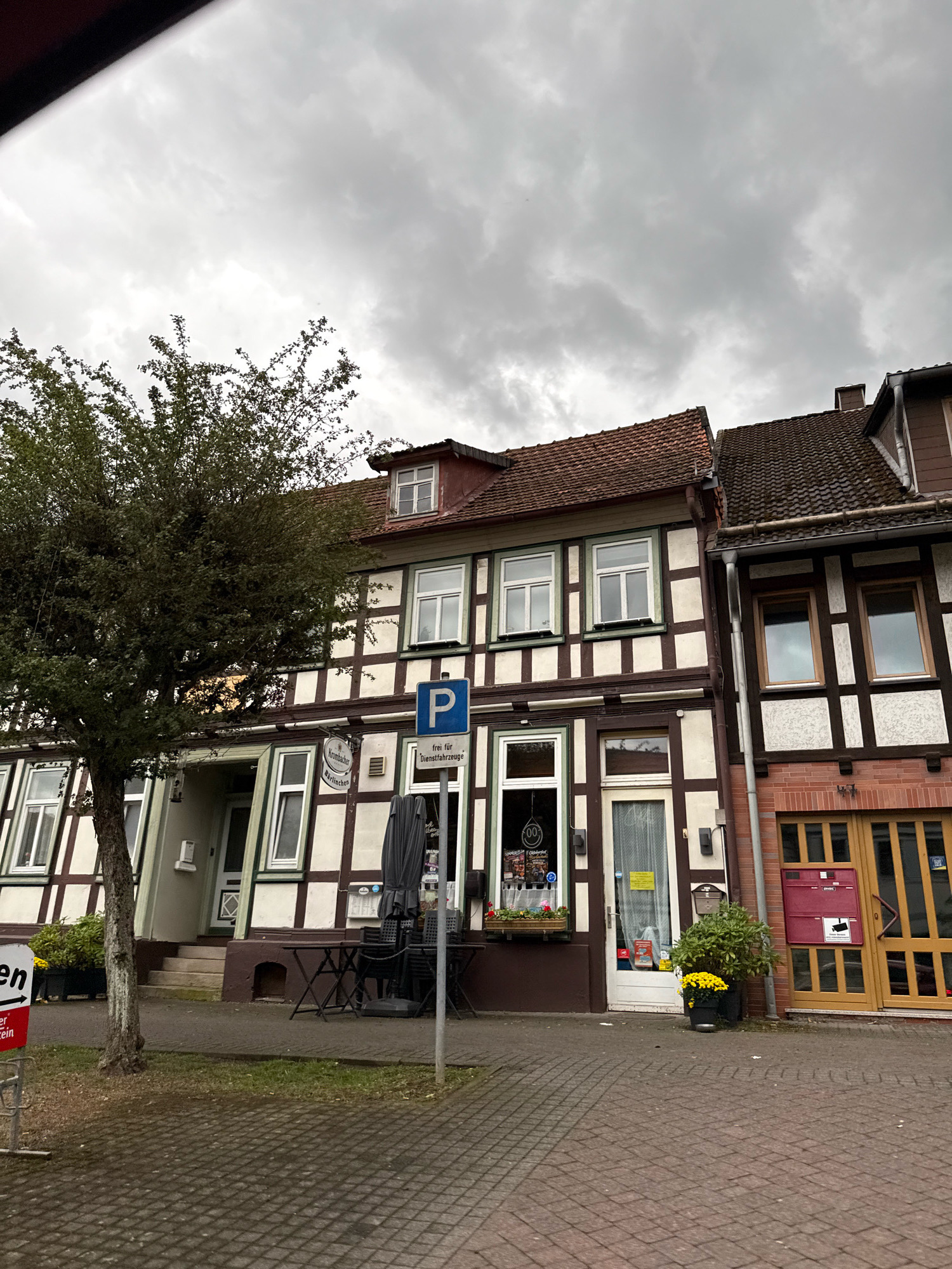 The image shows a cloudy day in Bad Sachsa, Germany. The street scene features traditional half-timbered buildings with brown and cream facades. A large, leafy green tree stands in front of one of the buildings on the left, with its branches extending towards the top of the frame. A paved walkway runs in front of the buildings, and a brick-paved road is visible in the foreground.
The building on the left has multiple windows, some with white frames and others with green trim. A sign above an entrance reads "Krombacher Bügellos". An outdoor seating area with tables and chairs is set up in front of the establishment, partially covered by a dark umbrella. A blue parking sign with a white 'P' is prominently displayed on a pole.
To the right of the first building is another similar structure, also with half-timbered details. This building has brown window frames. Further to the right, a brick facade building with a large wooden-framed glass door is visible. A red mailbox with a security camera sticker is mounted on this building. Yellow potted flowers add a splash of color to the scene, placed near the entrance of the second building. The sky is overcast with heavy grey clouds.