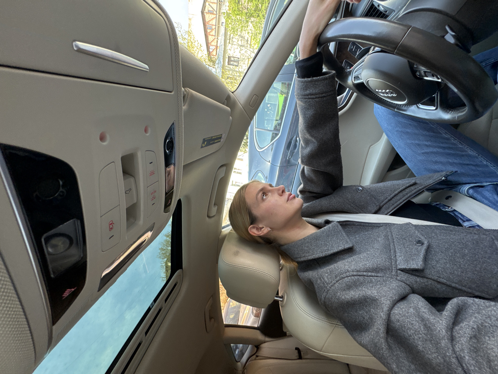 A woman with blonde hair is sitting in the driver's seat of a car, resting her head on the headrest and looking upwards. She is wearing a grey jacket and blue jeans. Her right hand is on the steering wheel. The car has a sunroof. The car appears to be parked on a street in Tbilisi, Georgia, based on the architectural style of the buildings visible through the car windows. There is another car parked alongside the vehicle.