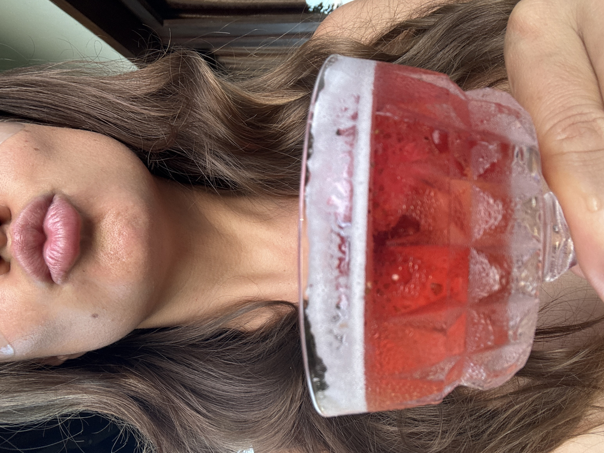 The image shows a person from the neck up, with wavy brown hair, fair skin, and full lips. A clear glass filled with a red liquid and condensation is held adjacent to their face. The drink appears to be a beverage, possibly a cocktail, with a geometric ice cube pattern. The subject is not exhibiting a clear action or interaction, but rather posing or holding the glass. The background is blurred, suggesting an indoor environment. There is no information in the provided image about the location being in the city of Kuta Selatan, Indonesia.