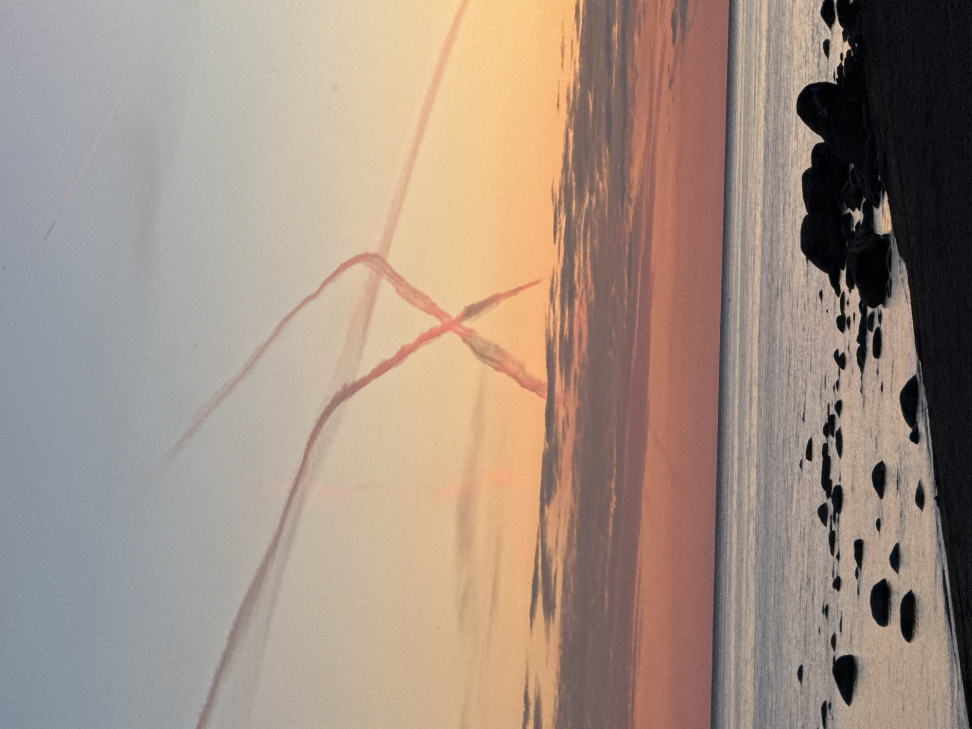 The image depicts a sky filled with contrails, likely from an airshow, against a sunset backdrop. The contrails form a prominent "X" shape in the upper portion of the sky, with additional trails extending upwards and to the left. The sky transitions from a pale blue at the top to hues of orange and pink near the horizon. Below the sky is the calm sea, reflecting the colors of the sunset. The foreground shows a rocky shoreline with dark, rounded stones scattered on the beach and in the shallow water. The beach itself appears dark and sandy. The overall scene is a serene coastal landscape at dusk, with the striking visual of the contrails adding a dynamic element. The location is identified as Zelenaya Roshcha, Russia.