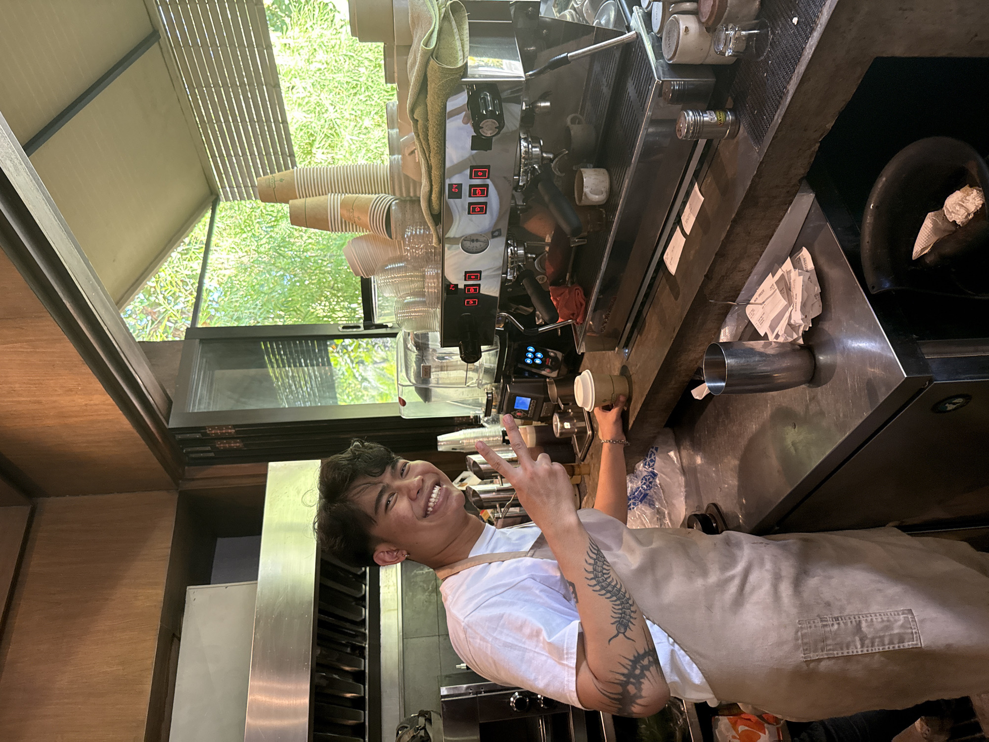 An indoor scene in a coffee shop in Kuta Selatan, Indonesia, shows a smiling barista posing behind a professional espresso machine. The barista, with dark hair and a bright, toothy smile, is making a peace sign with their right hand while holding a disposable cup with their left. They wear a white t-shirt and a light-colored apron, and their left arm features extensive tattoos, including a prominent centipede-like design and a spiky tribal pattern.
Behind the barista is a large, polished stainless steel espresso machine with multiple spouts, various buttons (some illuminated red and labeled 'P', 'C', 'D'), a pressure gauge, and a digital display. Stacks of paper and clear plastic cups are arranged on and around the machine. The dark counter in front of the barista holds small containers, a metal shaker with "SAH" visible, and a stack of paper receipts, one partially legible as "NEW ORDER". A black bowl with crumpled paper is also on the counter.
The setting appears to be an open-air or well-lit space, with large windows or openings revealing lush green foliage outside, indicating a daytime, natural, and possibly tropical environment. A wooden ceiling and a slatted awning structure are visible. The overall mood conveyed by the barista's expression is friendly and welcoming.