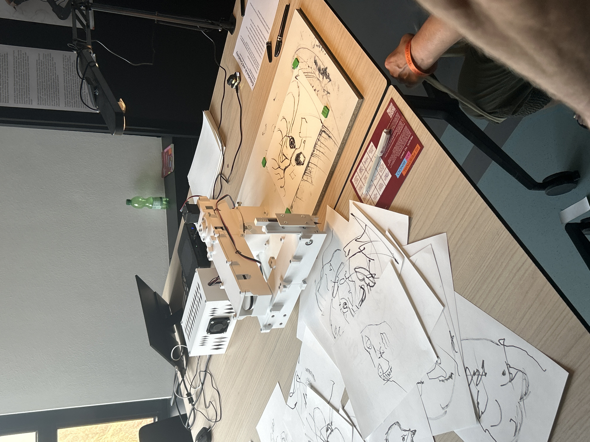 The image depicts a close-up, high-angle shot of a table with various items for what appears to be an art or technology project. In the foreground, scattered papers with abstract sketches are visible. To the right, a person's arm and wrist, adorned with an orange wristband, are resting on the table. The table surface is light-colored wood.
Dominating the mid-ground is a white robotic arm assembly mounted on a white base, positioned over a wooden board. The board itself has a sketch of a face on it, secured by small green objects. To the left of the robot, a white box with vents and a fan is present, along with a laptop and cables. Further back, a clear plastic bottle of liquid rests on a dark surface, next to a stack of papers.
In the background, a large, dark poster or screen with text is visible. The environment suggests an indoor setting, possibly a workshop, exhibition, or classroom, with neutral-colored walls. Lighting appears to be coming from overhead and possibly from a lamp mounted on a black arm in the upper right portion of the image. The overall impression is one of active creation or demonstration.