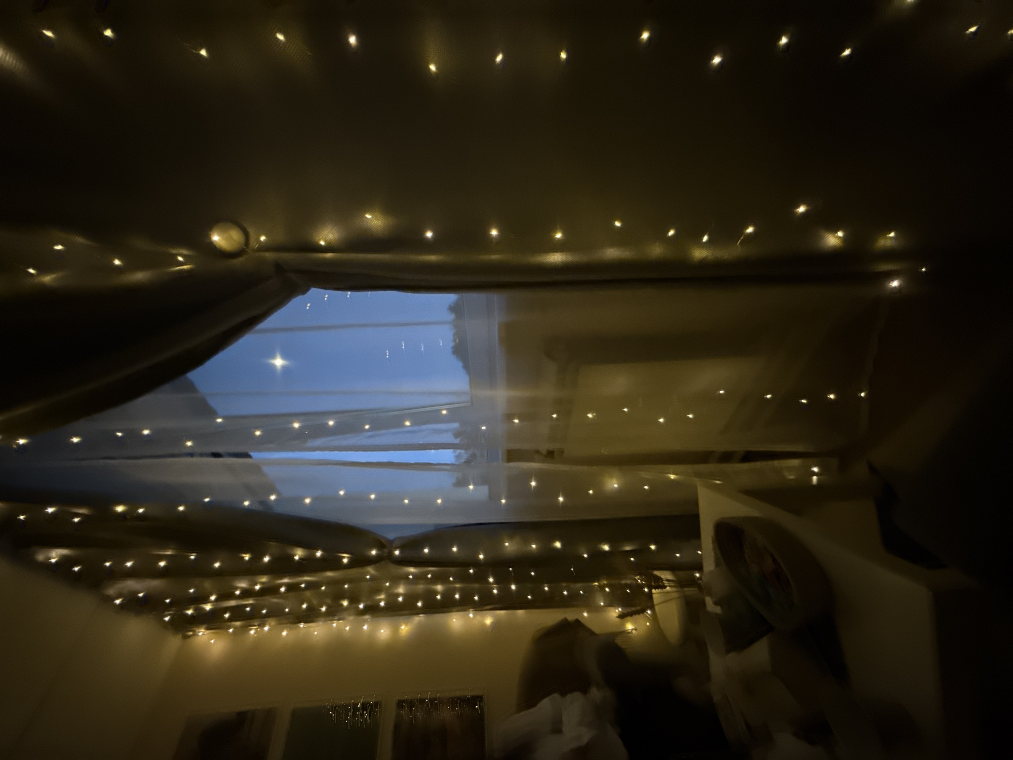 The image depicts an indoor scene, likely a bedroom or similar space, decorated with strings of fairy lights. The lights are strung across the ceiling and around a structure resembling a draped canopy or bed frame, creating a soft, warm glow. A window is visible through the draped fabric, revealing a twilight sky. Furniture is partially visible in the foreground, including what appears to be a bed with rumpled bedding. The overall impression is that of a cozy, dimly lit space. There is no definitive evidence to confirm the location is Chaikovsky, Russia.