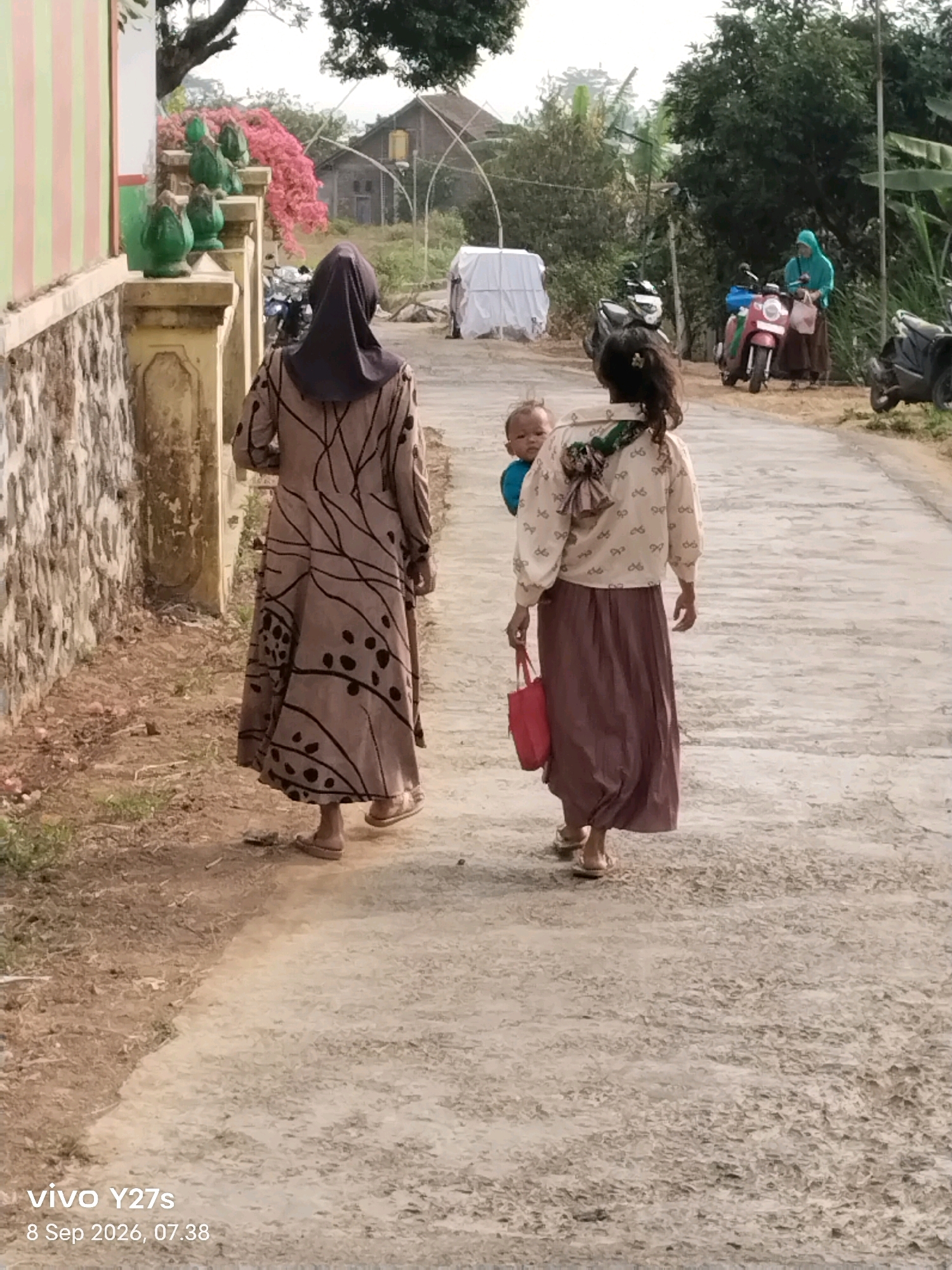 In the city of Tamansari, Indonesia, a woman wearing a brown hijab and a patterned dress walks along a dusty path. A child is strapped to her back. To her right, another woman in a light-colored blouse and a long brown skirt, also carrying a child on her back, walks ahead. The child, visible over the woman's shoulder, looks towards the camera. In the background, several motorcycles are parked. A woman in a blue hijab stands near one of the motorcycles. The environment appears to be a rural or suburban area, with some vegetation and a building visible in the distance. The overall scene depicts people on a casual stroll.