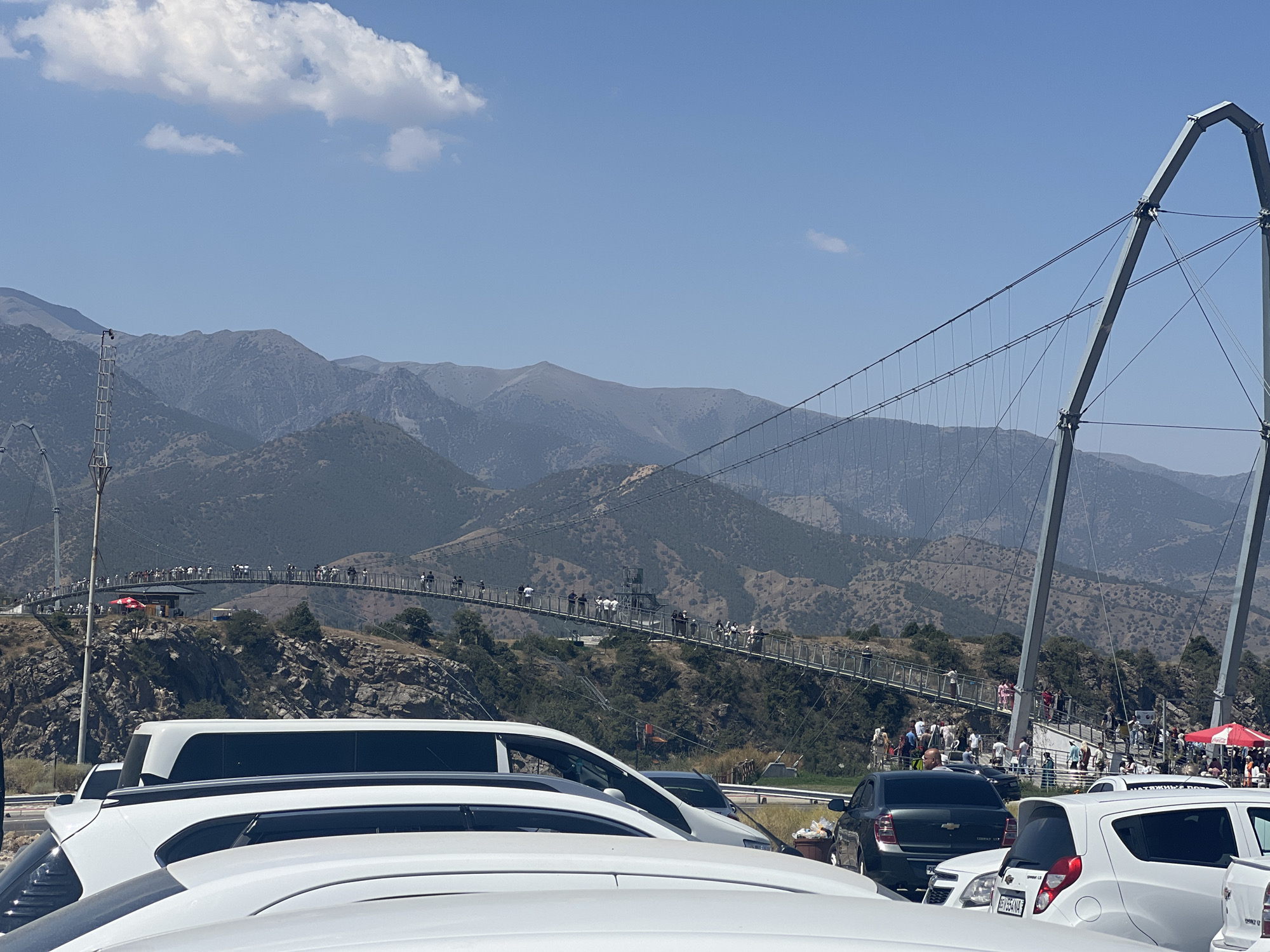 The image depicts a sunny day with a clear blue sky and scattered clouds, above a mountainous landscape. In the foreground, several white vehicles are parked, partially obscuring the view. A long, modern suspension bridge, filled with numerous people, stretches across a valley in the middle ground. The bridge is supported by large metal structures. The surrounding terrain is rugged and dry, with sparse vegetation on the hillsides. The location is identified as the city of Dardar, Uzbekistan.