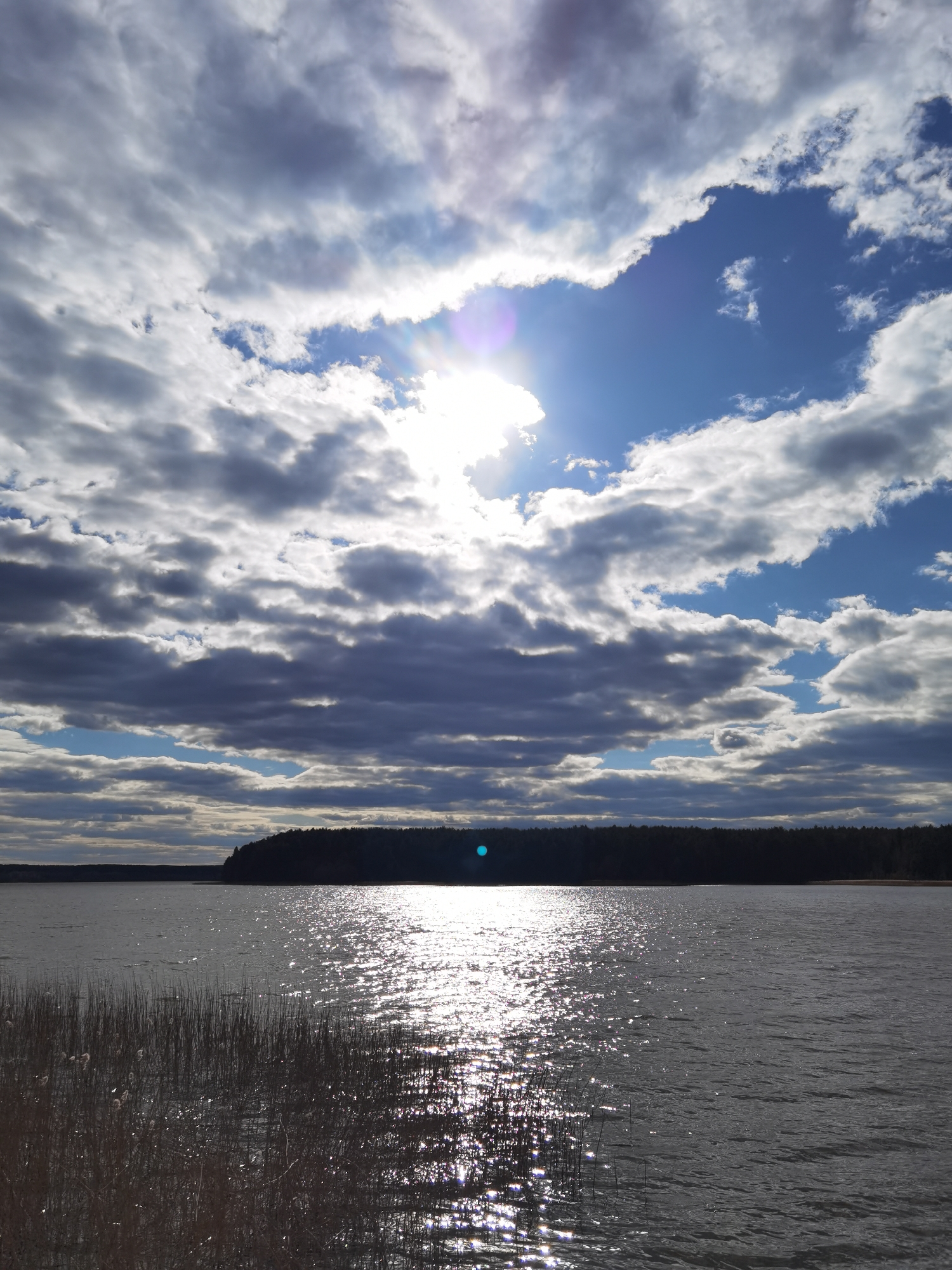 The image depicts a body of water, likely a lake or a wide river, reflecting sunlight. The sky is partially obscured by clouds, with a patch of blue sky visible. The sun is shining through the clouds, creating a bright glare. A dark tree line is visible in the background across the water. Some reeds or tall grasses are growing in the foreground on the left. Based on the location information, this is likely the Votkinsk Reservoir or a similar body of water near Votkinsk, Russia. There are no people visible in the image. The scene is a natural landscape with water, sky, clouds, trees, and vegetation.