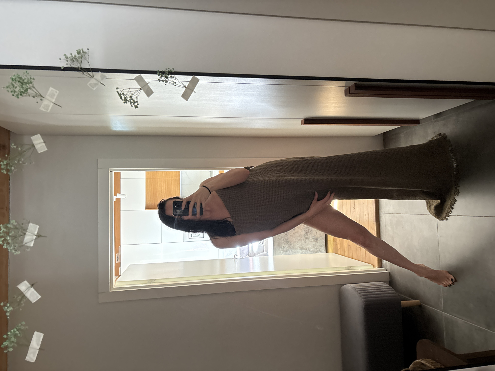 A mirror selfie shows a woman in a room in Chaikovsky, Russia. She holds a phone in her left hand, partially obscuring her face. She is draped in a brown, fringed textile, exposing one leg. Behind her is an open doorway leading to a bathroom with white tiles and wooden accents. The room also contains a gray upholstered bench. The walls are light-colored, and there are decorative flowers taped to the surface of the mirror.