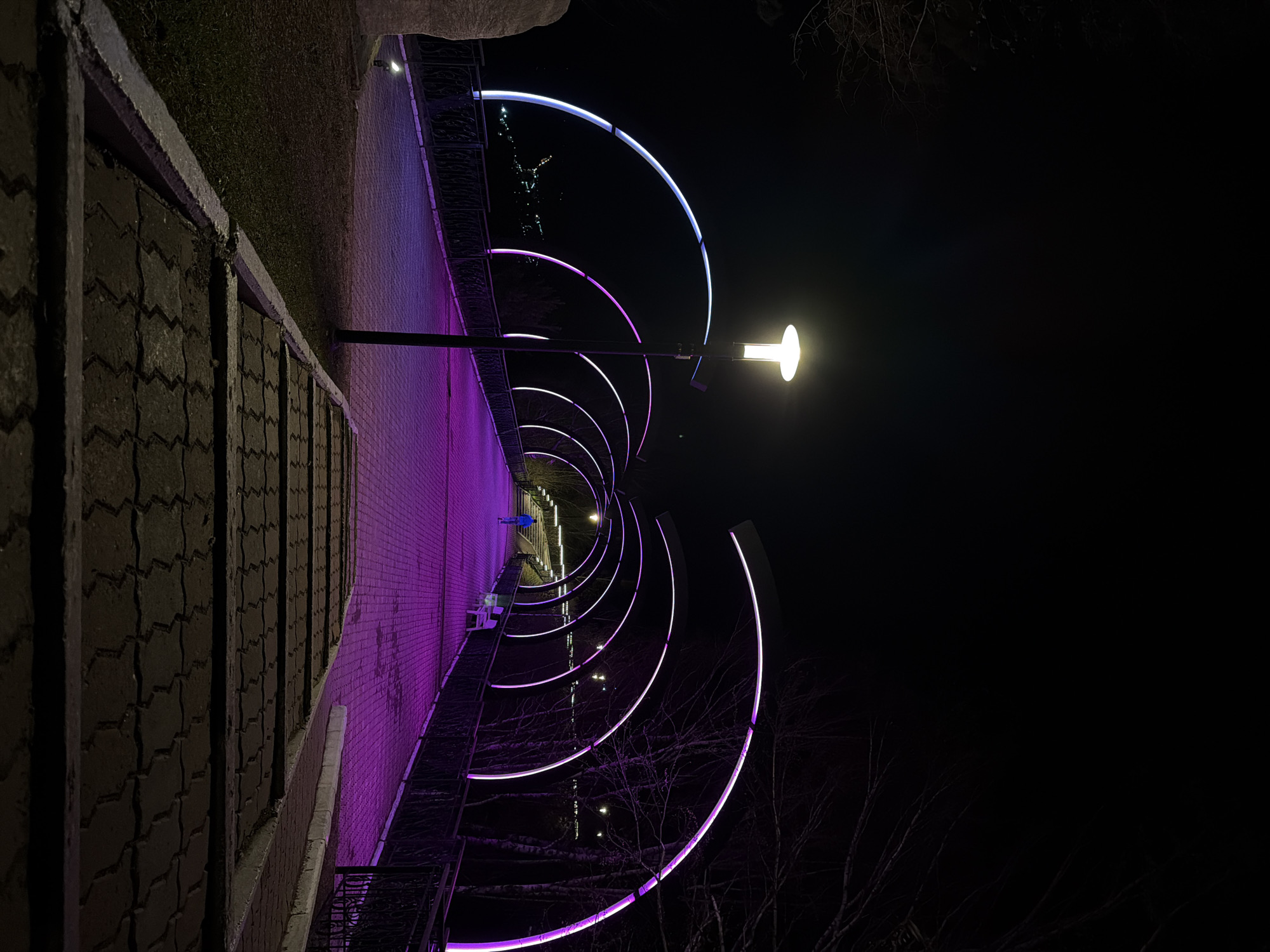 The image shows a pedestrian walkway at night in Слизнево, Russia. The walkway is illuminated by a series of curved, neon-colored light installations, progressing from white to purple. A traditional streetlight is also visible, providing a warm, yellow light. A person in a blue jacket is walking along the walkway in the distance. The walkway is lined with a low brick wall on one side and a grassy area on the other. Bare tree branches are visible against the dark sky.