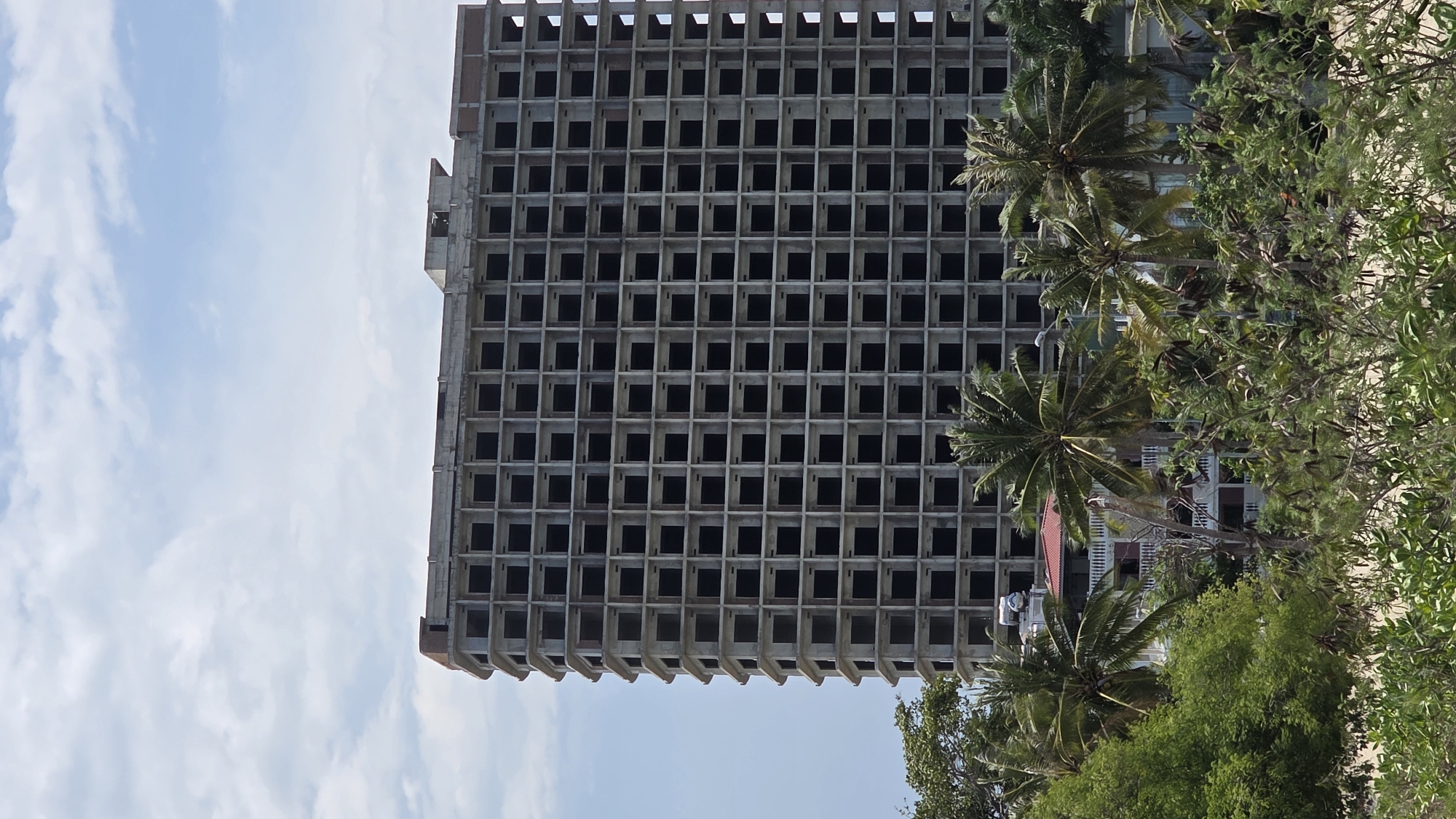 The image shows an unfinished concrete building with numerous window openings, located in Nam Nha Trang Ward, Vietnam. Palm trees and other greenery surround the building. The sky is partially cloudy. A smaller building with a red roof is visible near the base of the unfinished structure. A person wearing white is seen on or near the smaller building.