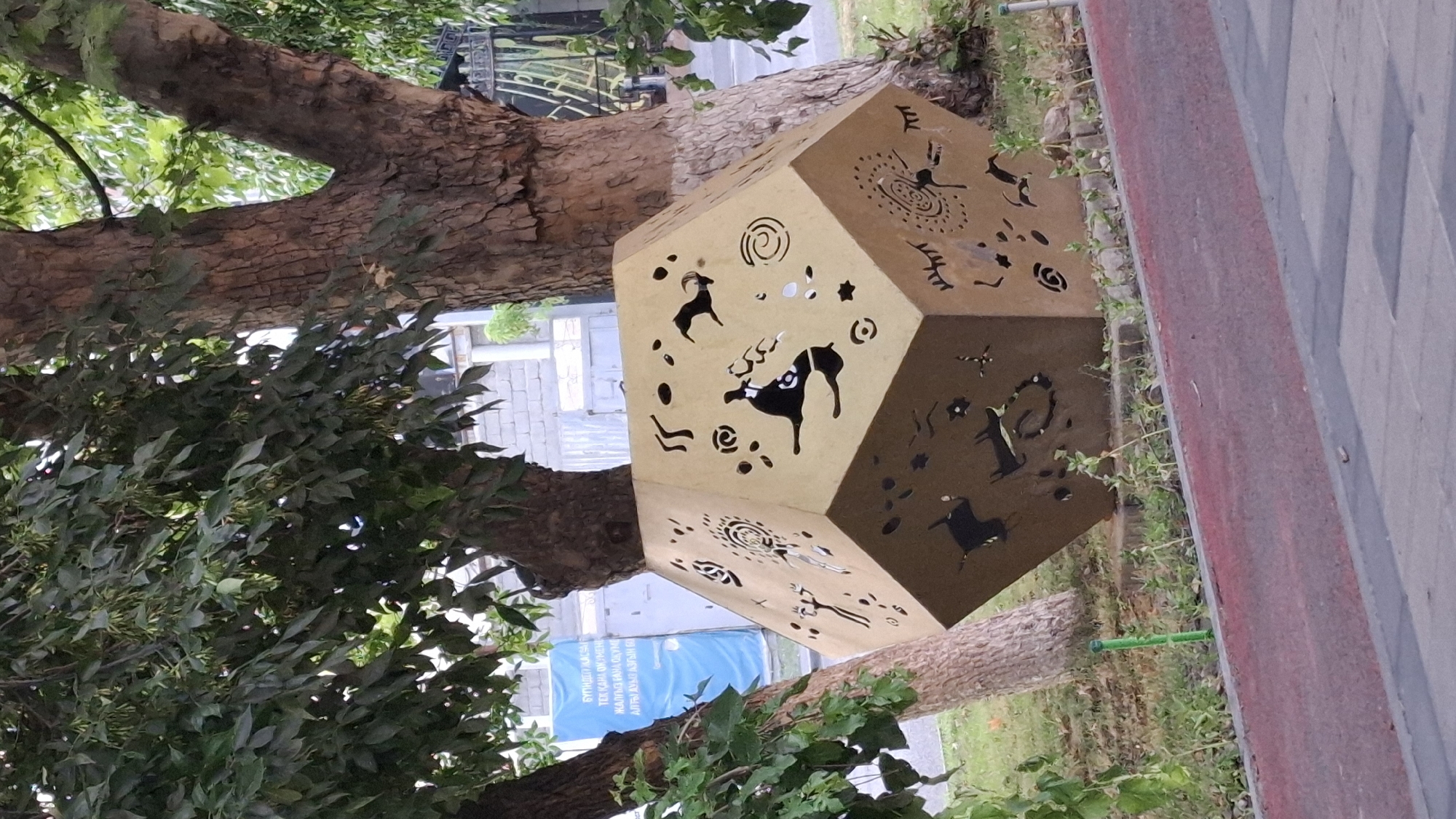 In the city of Shymkent, Kazakhstan, a polyhedral sculpture with cutouts depicting ancient petroglyphs is displayed outdoors amidst trees. The sculpture, made of a light-colored material, features geometric shapes and stylized animal and human figures. In the background, a blue banner with text is visible, along with a brick wall and a metal fence. The area appears to be a public space, possibly a park or a street side, with a red-colored pedestrian path in the foreground. The lighting suggests it is daytime, with shadows cast by the trees and the sculpture.