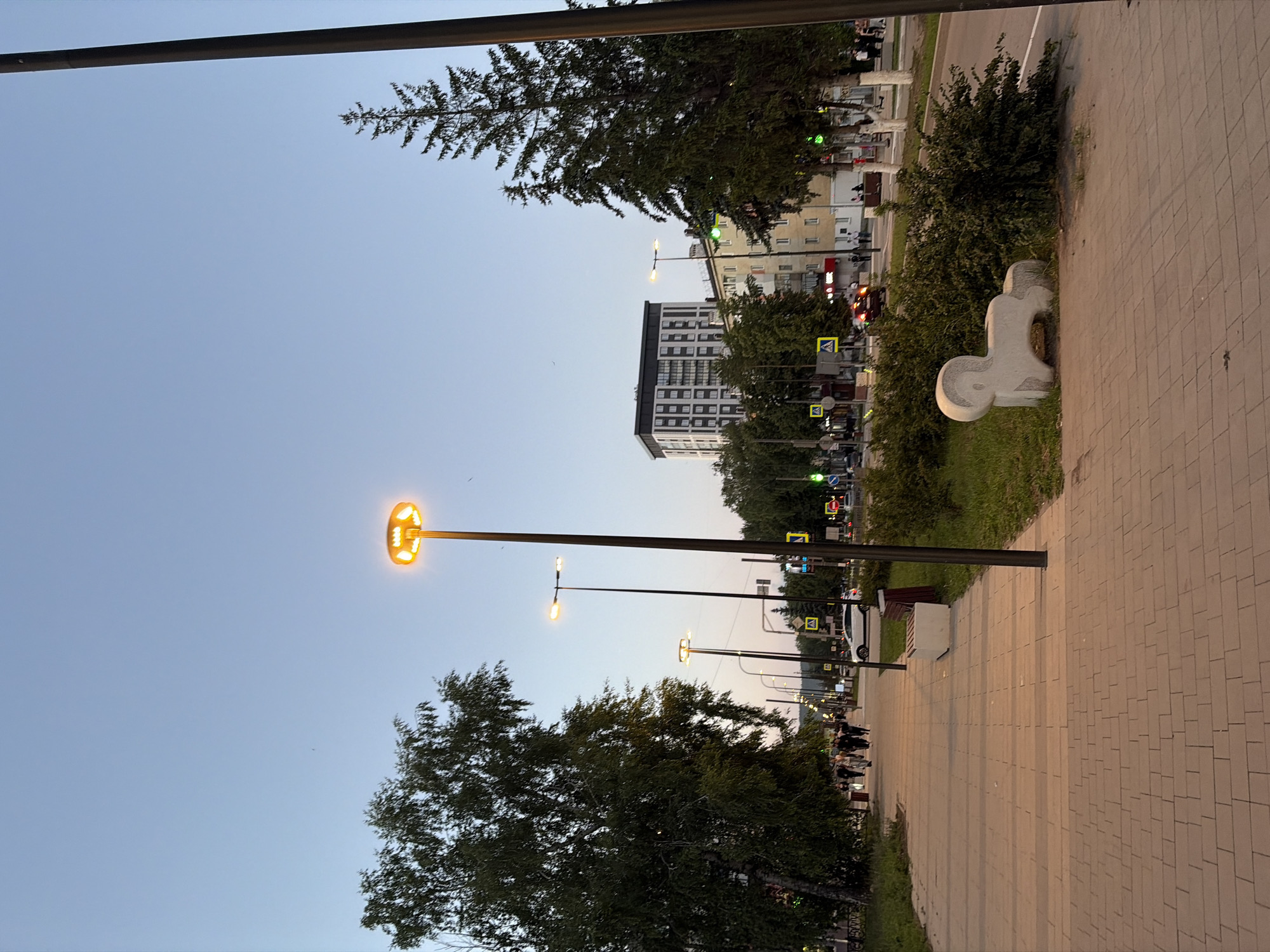 The image depicts an outdoor urban scene at dusk, likely in the city of Yarykovo, Russia. A paved pedestrian walkway runs along the left side of the frame, leading towards a street with vehicular traffic. Several streetlights illuminate the walkway, and a modern building is visible in the background, along with other residential and commercial structures. People are walking along the path, and cars are present on the street. Trees line the walkway and the street, contributing to the urban greenery. A notable object on the grass verge is a stone sculpture resembling a stylized animal, possibly a ram. The sky is a clear, light blue, indicating the transition from day to night.