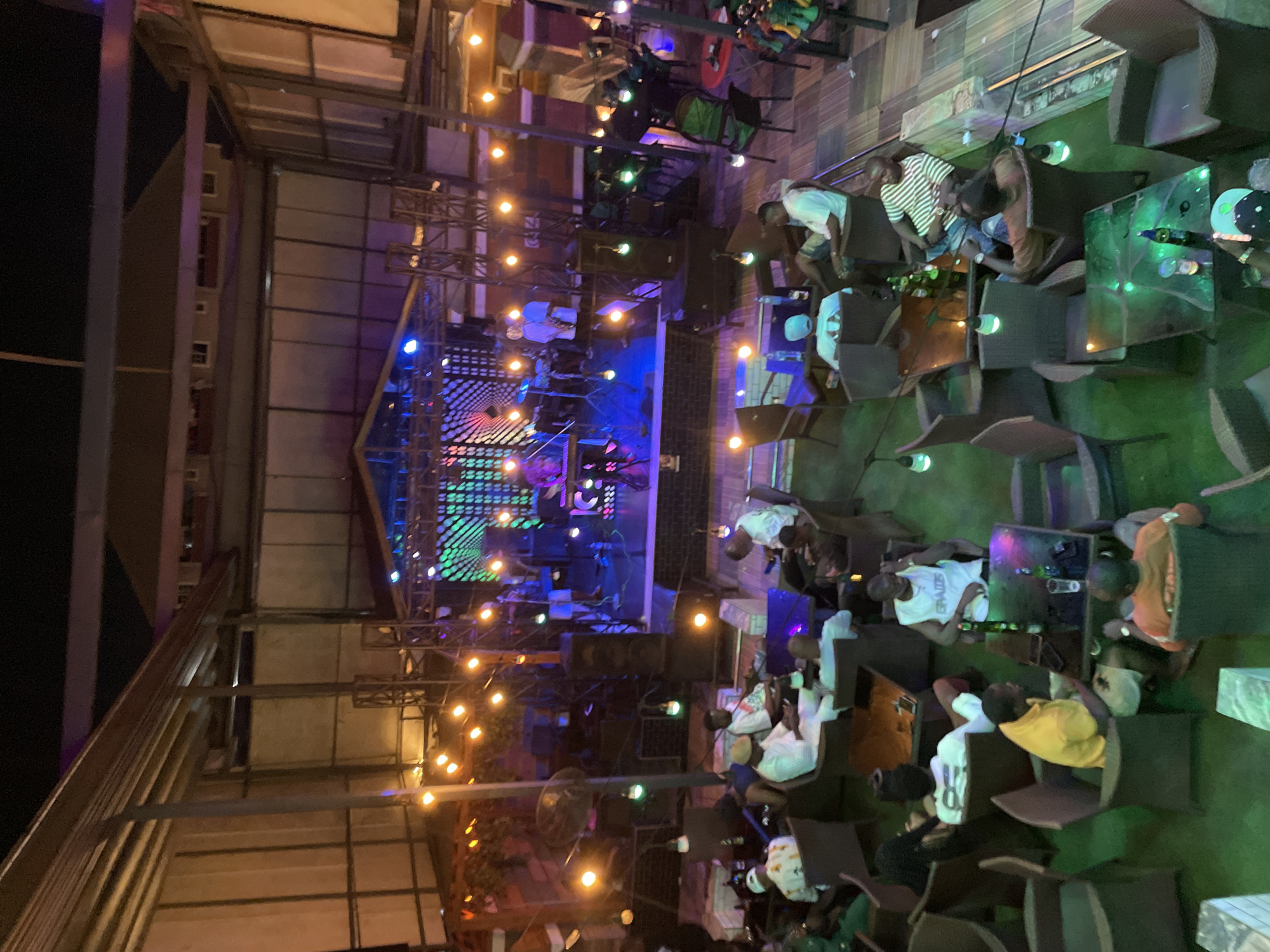 The image shows an outdoor venue in Obosi, Nigeria, likely a bar or entertainment space at night. A stage is set up with lighting and sound equipment, suggesting a performance or event is taking place or about to. Numerous people are seated at tables, some appearing to be in conversation, while others seem to be watching the stage. Tables are set with drinks and personal items. The venue is illuminated with overhead string lights and stage lighting, with the architecture of the building visible in the background.
