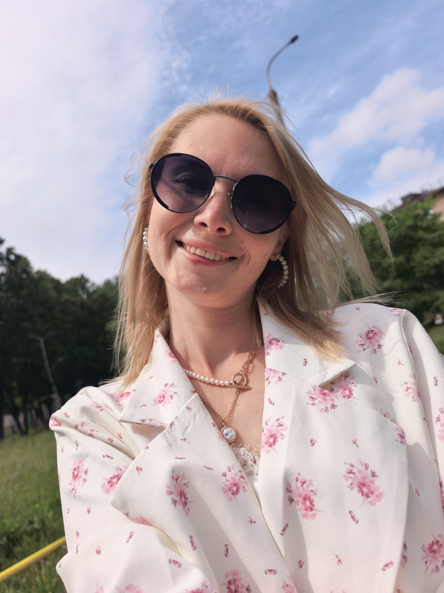 The image is an upward-angled, well-lit selfie of a smiling woman outdoors in Sterlitamak, Russia. The woman, with blonde or light-colored hair appearing to be gently wind-blown, is wearing large, round, dark sunglasses that reflect the bright sky. She smiles broadly, looking directly at the camera, conveying a cheerful and happy mood.
She is dressed in a white blazer or jacket adorned with a delicate pink floral pattern, with a white, lace-trimmed top visible underneath. Her accessories include elegant pearl hoop earrings and a layered necklace featuring a strand of pearls, a gold chain with a circular pendant, and another gold chain with a T-bar clasp.
The background depicts a clear, sunny day with a vibrant blue sky and scattered white clouds. Lush green trees and grass are visible, suggesting a park or a natural outdoor setting. A dark, curved street lamp or utility pole extends into the sky on the right, and a faint hint of a distant building is also visible. A yellow railing or bar can be seen in the bottom left corner, indicating a public walkway or area. There is no visible text in the image.