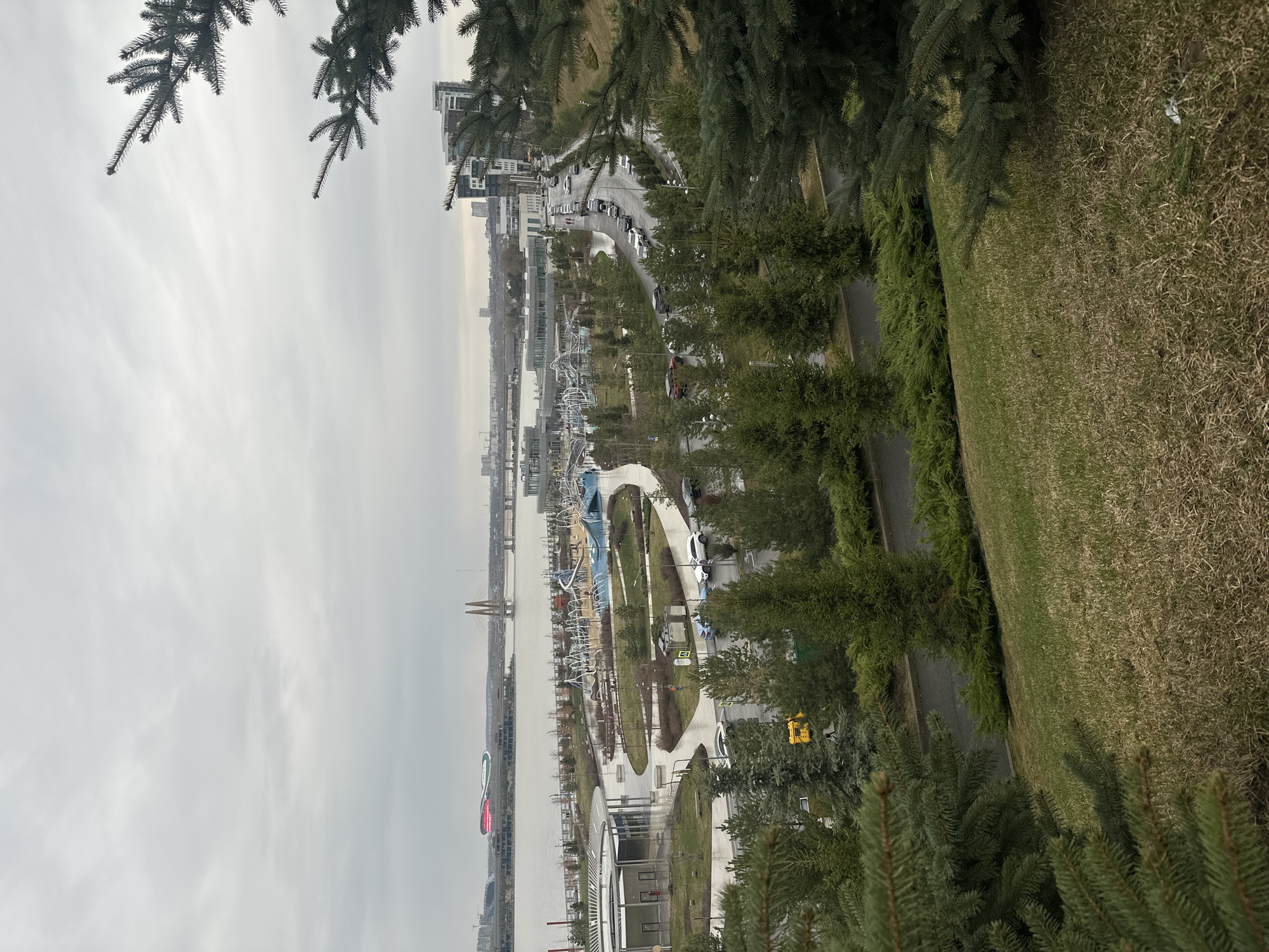 The image depicts a panoramic view of Kazan, Russia, overlooking the Kazanka River and its surrounding urban landscape. From an elevated vantage point, possibly a hillside park, the foreground is dominated by evergreen trees and grassy terrain. The middle ground features a pedestrian walkway and a row of trees, paralleled by a road with parked cars. A playground with blue and white structures is also visible. In the distance, the river is spanned by cable-stayed bridges. Buildings and urban infrastructure form the backdrop against an overcast sky. No people are clearly visible. The general atmosphere is one of urban parkland adjacent to a riverfront development.