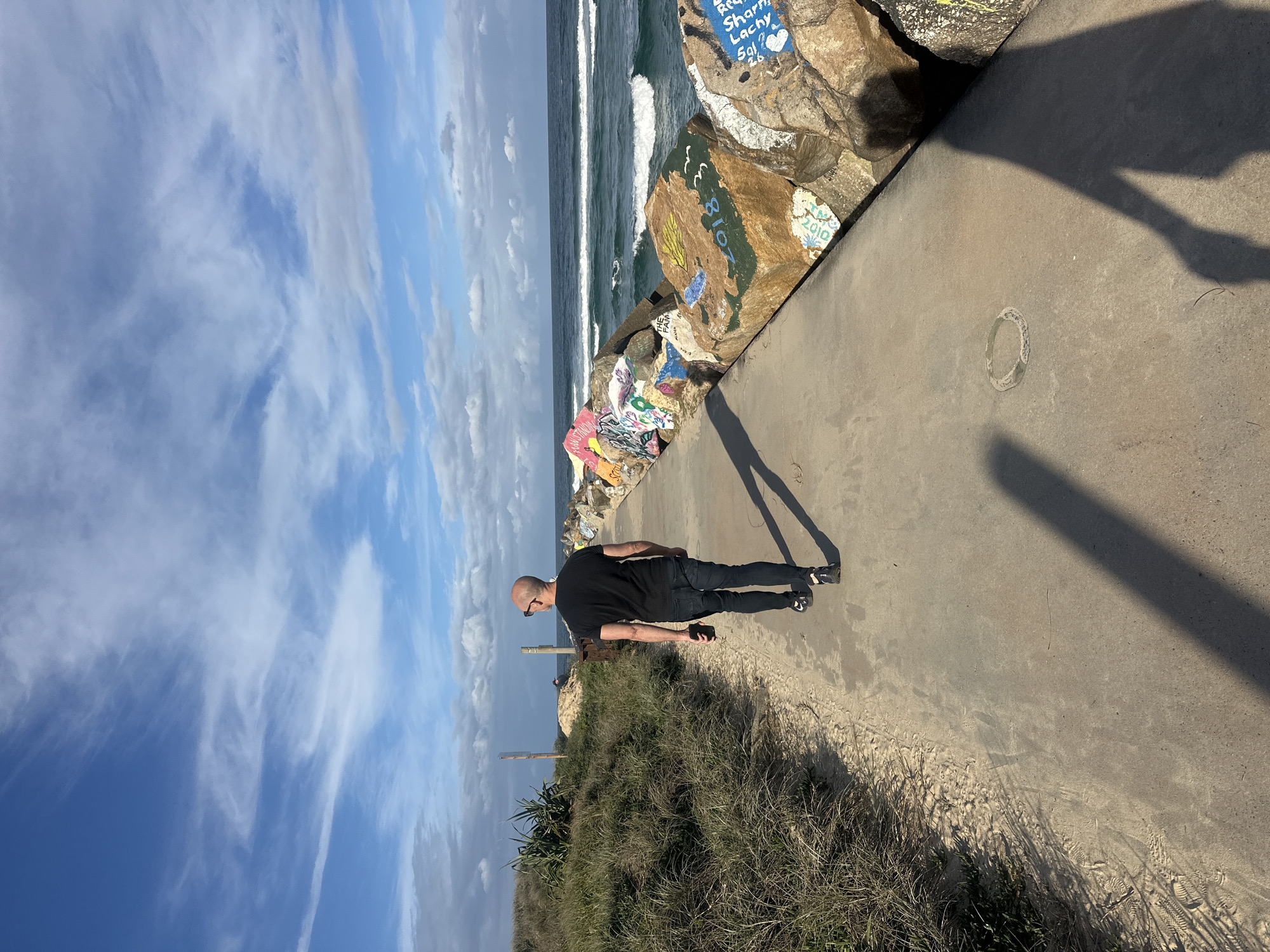 A bald man wearing sunglasses, a black t-shirt, and black jeans walks away from the camera on a paved path beside a rocky breakwater. The breakwater is covered in colorful graffiti. The ocean is visible to the right of the path, with waves breaking. To the left of the path is a sandy area with tall grass. The sky is blue with scattered white clouds. The scene appears to be a coastal path in Nambucca Heads, Australia.