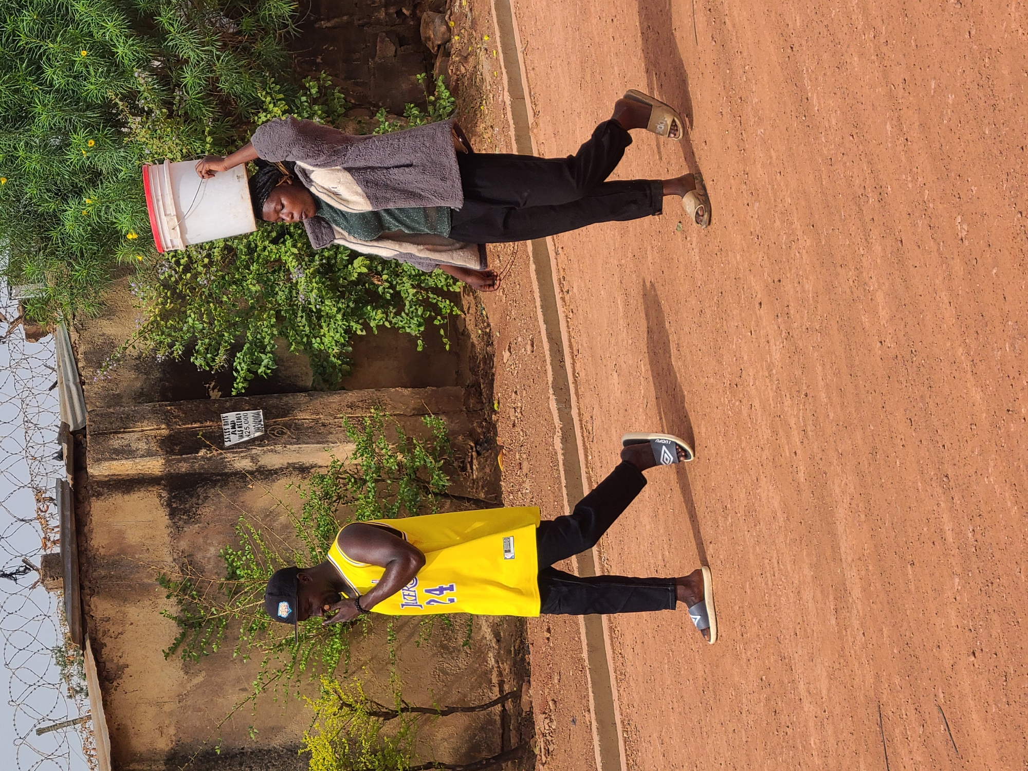This outdoor image, taken in Jos, Nigeria, depicts two individuals on a reddish-brown dirt road under bright daylight.
In the foreground on the left, a person, likely female, walks with a white plastic bucket with a red rim balanced on her head, likely transporting water. She wears a dark, possibly grey, patterned jacket or cardigan over a green top and dark trousers, with light-colored slide sandals. Her braided hair is visible.
Further back and to the right, another person, likely male, stands wearing a bright yellow basketball jersey with the number "24" and "LAKERS" (partially visible) on the front. He has on black pants, a black baseball cap, and dark slide sandals with white soles, marked "LUCKY" or "LUDWIG" on the strap. He appears to be looking downwards.
The setting includes lush green bushes and foliage on the left side of the frame, bordering a weathered concrete or stone wall. A barbed wire fence is visible on the far left, extending along the top of the wall.
On the wall behind the person in the yellow jersey, a white sign with black text is visible. The readable text on the sign includes: "SALES POINT", "A HALL", "NO ENTRY", "N25,000", and "UNAUTHORISED PERSON(S) ARE NOT PERMITTED". The "N25,000" likely refers to Nigerian Naira, indicating a price.
The strong shadows cast by the individuals and foliage suggest a sunny day, either mid-morning or late afternoon. The overall scene reflects daily life in an unpaved area of Jos, Nigeria.