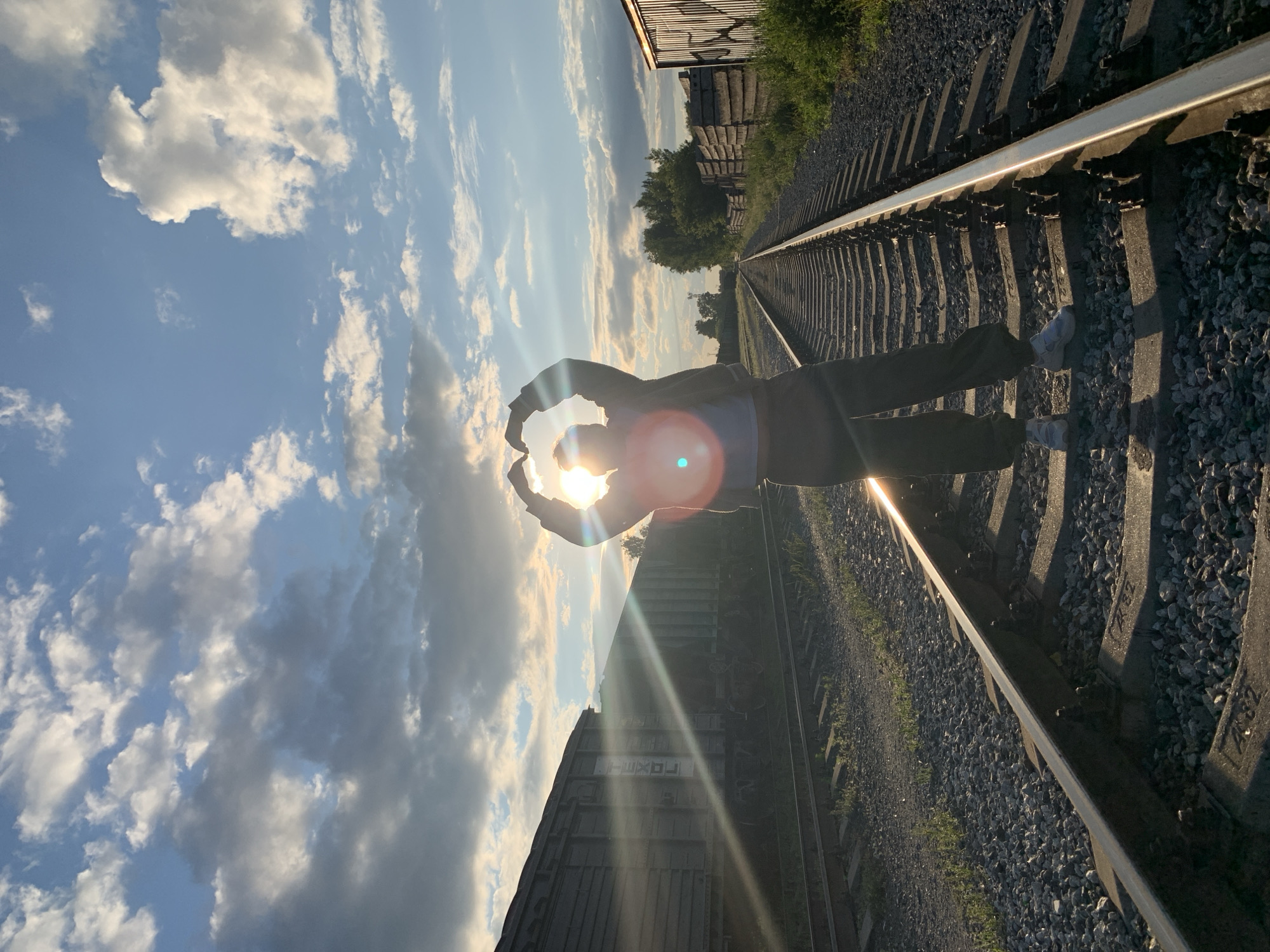 A person is silhouetted on railroad tracks, facing the camera and forming a heart shape with their hands above their head. The sun is setting behind them, creating a lens flare. The scene appears to be a disused railway yard on the outskirts of Chaikovsky, Russia, as indicated by the abandoned freight cars and weathered wooden structures. The sky is cloudy, with patches of blue peeking through. The overall mood is peaceful and nostalgic.