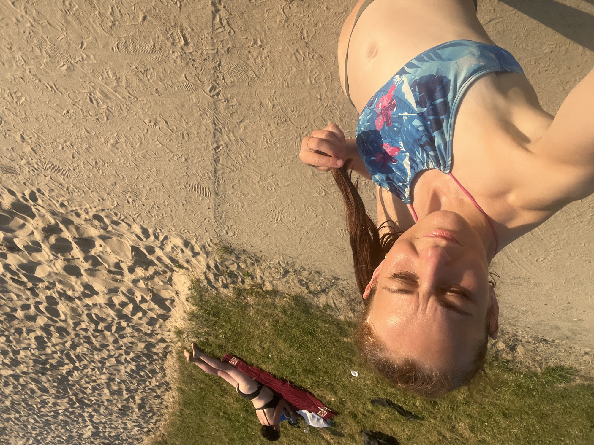 An outdoor, eye-level, wide shot shows two women on a sandy beach with a grassy embankment. The woman in the foreground is in the lower left corner, looking down and to her left. She is wearing a blue and pink floral bikini top and has her hair pulled back in a ponytail. She is holding a strand of her hair with her right hand. The second woman is further back on the grassy embankment, lying on her stomach on a red towel. She is wearing a black bikini and looking down at a white object in front of her. The sand is imprinted with numerous footprints. The lighting suggests it is late afternoon or early evening. The overall environment is a public beach.