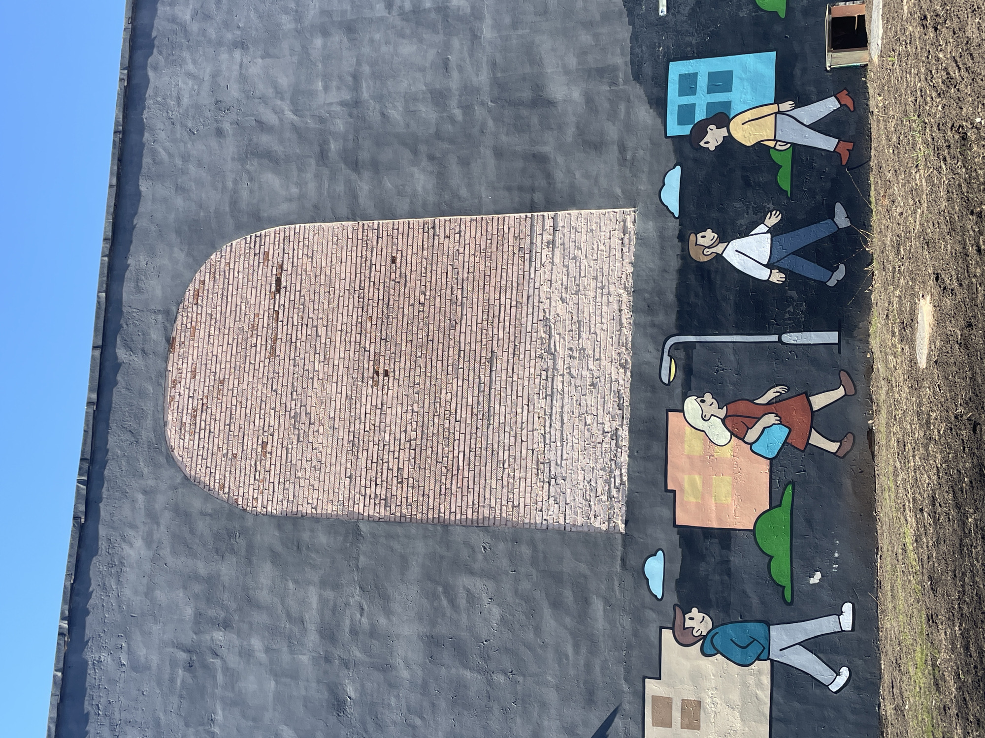 The image shows a building exterior in Izhevsk, Russia. A mural is painted on a grey wall. The mural depicts cartoon-like figures of people near buildings, clouds and a streetlight. There is an unpainted bricked section that forms a curved top, resembling a door or window. Dirt is along the top-right of the wall. The sky is visible in the upper-left.