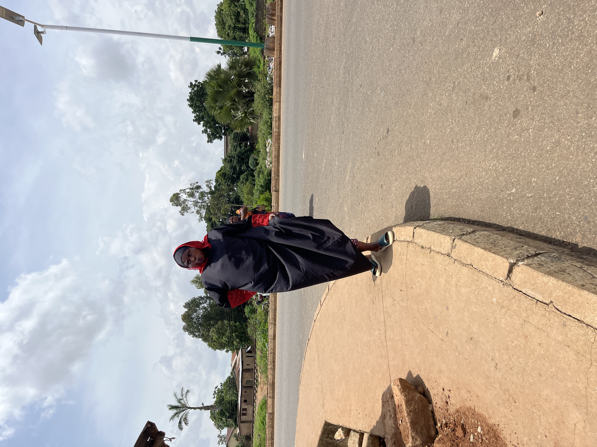 In Bukuru, Nigeria, a woman stands on a concrete sidewalk next to a paved road under a cloudy sky. She is wearing a black abaya with a red hijab and red embroidered sleeves. Her left hand is on her hip, and her right hand is visible to her side. She wears dark blue sandals with a light sole. The sidewalk is cracked and uneven, with loose dirt and rocks in the foreground. The road is asphalt and appears to be a two-lane road. In the background, there are trees, buildings, a lamppost with solar panels, and a railway track. A second person is partially visible behind the woman. The scene depicts a typical street view in the town.