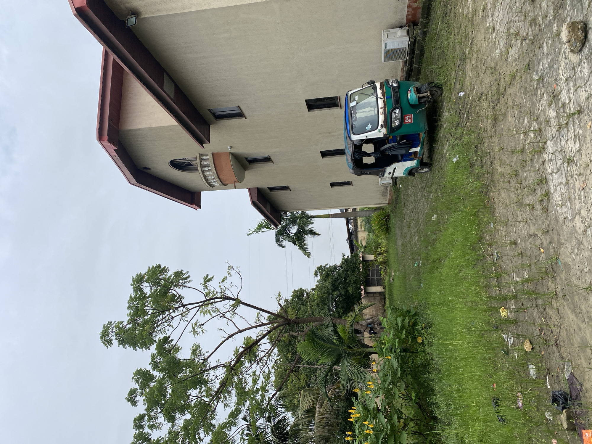 A three-wheeled auto rickshaw is parked on a paved surface outside a large, multi-story building with beige walls and a reddish-brown roof. The auto rickshaw is primarily white and green, with a blue trim. There are two air conditioning units mounted on the exterior wall of the building. The surrounding area is overgrown with green grass and some yellow flowering plants are visible to the left. A large tree with green leaves stands to the left of the building. The sky is overcast and gray.