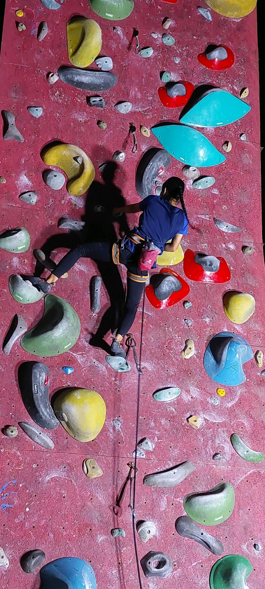 The image shows a person, likely a child, climbing a red climbing wall. The climber is wearing a blue t-shirt and black leggings, with a harness and climbing rope attached. They are in the middle of the wall, with their legs positioned on different climbing holds. The wall is covered with various colorful climbing holds of different shapes and sizes, including yellow, green, blue, and red ones. A shadow of the climber is cast on the wall. The environment appears to be an indoor climbing gym.