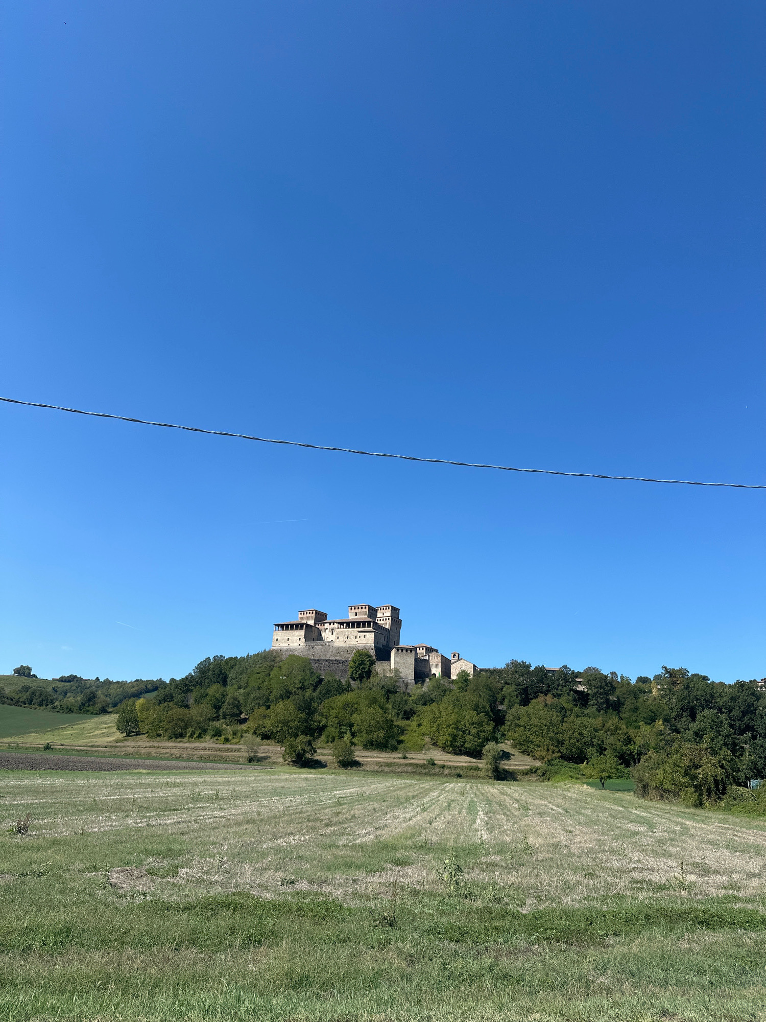 The image depicts a clear blue sky above a hill topped by a castle. The castle appears to be constructed of stone and has multiple towers and crenellated battlements. It is surrounded by dense green trees. In the foreground, there is a field with dry, cut grass, indicative of agricultural activity. A thin, dark wire runs horizontally across the upper portion of the image. The scene is set in a rural landscape. The castle is identified as the Castello di Torrechiara, located in the municipality of Langhirano, Italy.