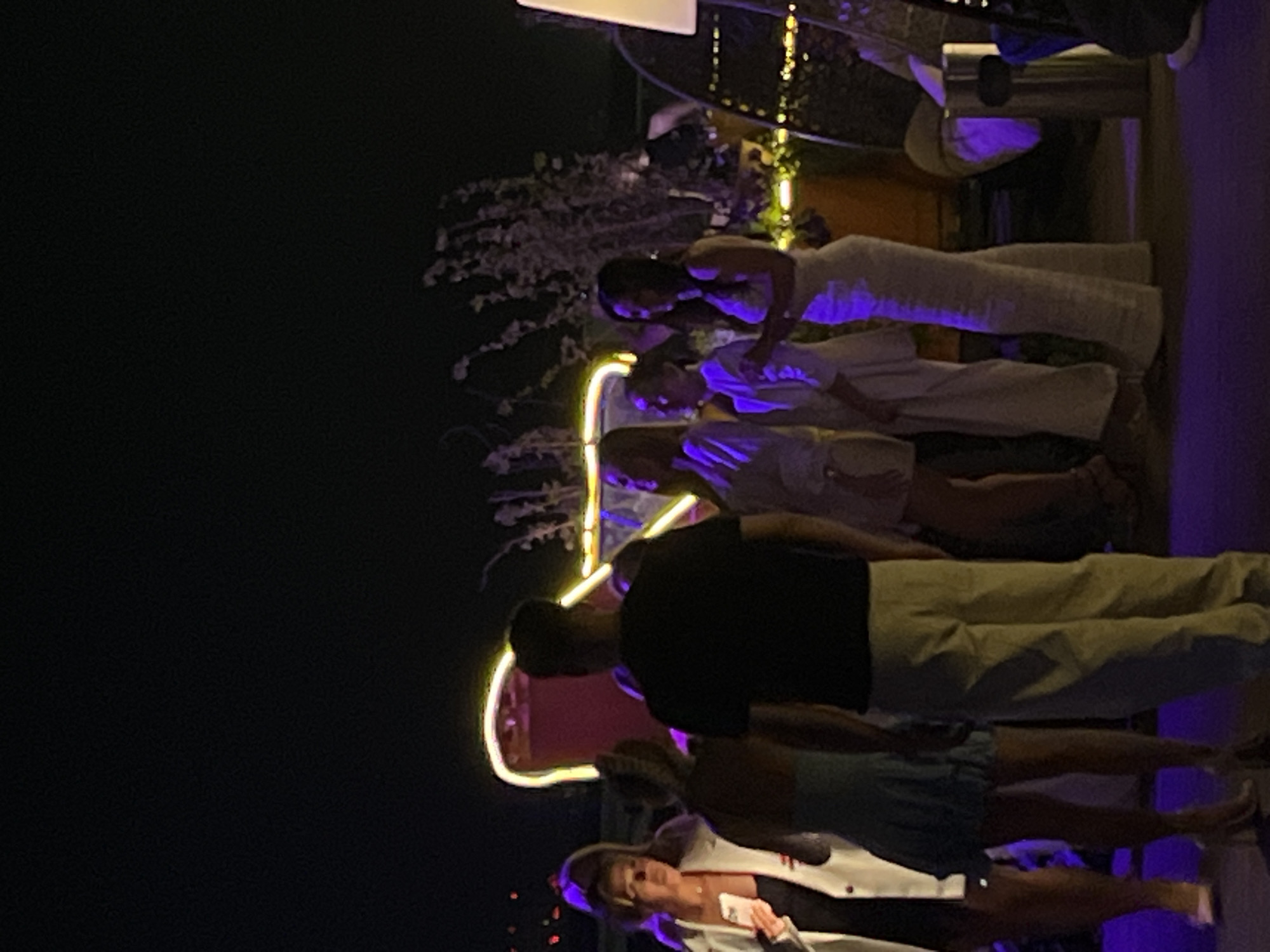 The image shows a group of people at night. They are gathered in what appears to be an outdoor setting, potentially a rooftop terrace, in Kazan, Russia. A neon sign is illuminated in the background. The group includes several women in dresses and lighter-colored outfits, and a man in a dark shirt and khaki pants. One woman in the foreground is wearing a white jacket. Some people are talking, but the overall activity is difficult to discern. The background is dark, with hints of foliage and a possible structure.