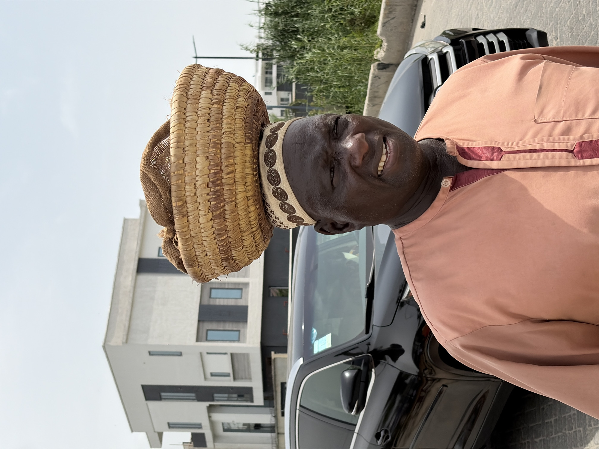 A man in Moba, Nigeria stands near a black vehicle and a modern building. He wears a peach-colored tunic with maroon trim and a woven straw hat. He is smiling. The location appears to be an outdoor urban setting.