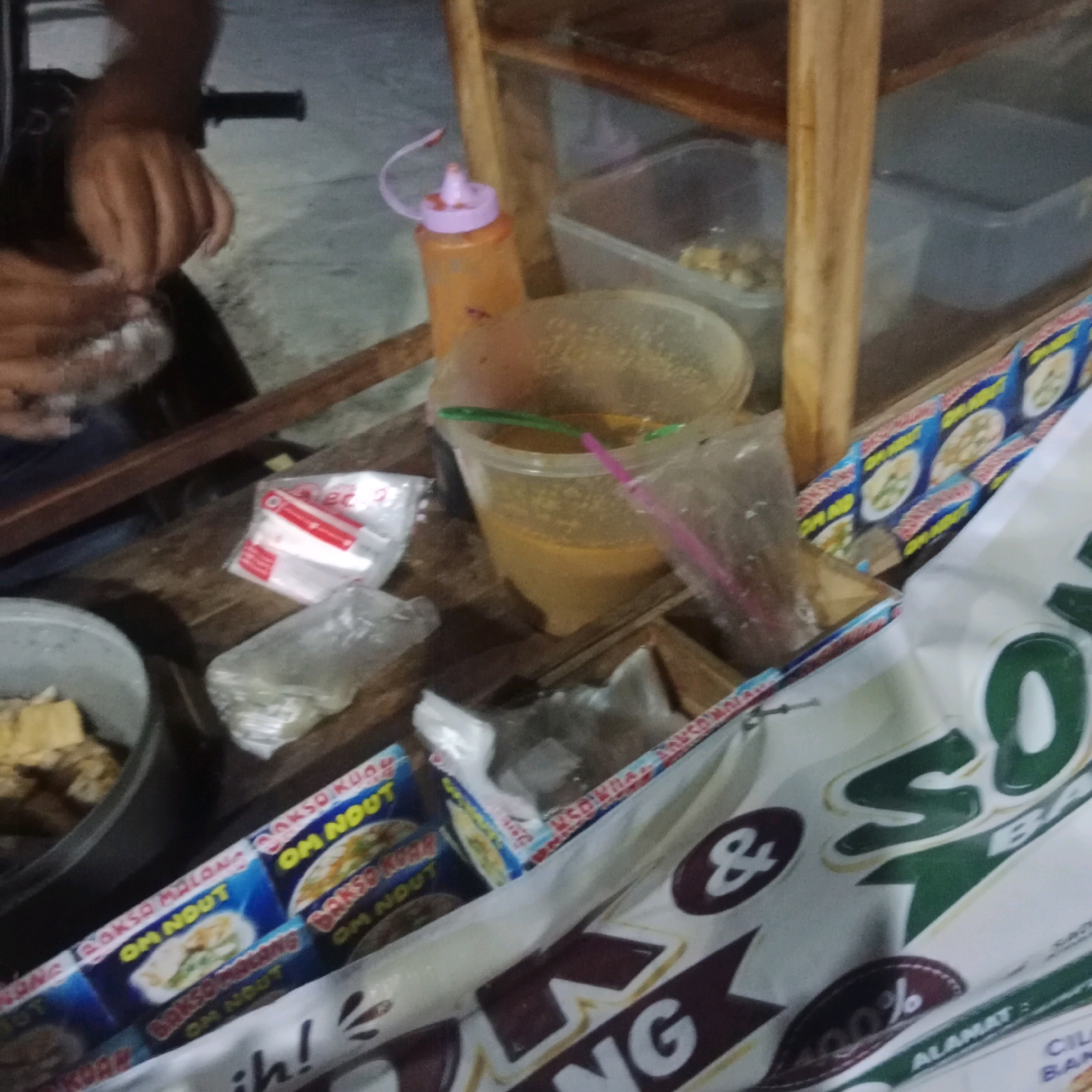 The image depicts a street food stall in Tlogorejo, Indonesia. The stall appears to be set up on a wooden cart, with various food items and supplies. In the foreground, there is a white plastic bag with large green lettering that reads "SO...". Next to it, there is another bag with a maroon circle containing a white ampersand symbol. Several packets of "BAKSO MALANG OM NDOUT" are visible, stacked and arranged. A person's hands are visible on the left, engaged in preparing or handling food.
Further back, a clear plastic container holds a yellowish liquid, with a pink straw submerged in it. A red and purple squeeze bottle, likely for sauce, is positioned near the container. The stall is constructed with wooden planks, and a transparent plastic material serves as a display or enclosure. The background is somewhat blurred, but suggests an outdoor, possibly urban, setting. The lighting is dim, suggesting it might be evening or nighttime.