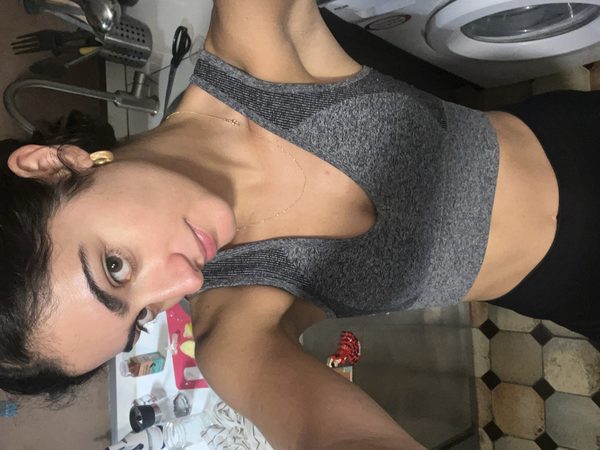 The media file captures a close-up, rotated portrait of a young woman within an indoor, domestic setting, likely in Paris, France. The woman has dark hair pulled back, prominent dark eyebrows, and a warm skin tone. She wears a grey, ribbed athletic crop top or sports bra and dark bottoms. A delicate gold chain necklace adorns her neck, and a simple gold earring is visible in her left ear. Her gaze is directed towards the camera with a subtle, calm expression, and her lips are slightly parted in a faint smile.
The background suggests a kitchen or utility area. To the left of her head, a metal kitchen faucet and sink are visible, along with a dish drying rack and what appears to be a pair of scissors hanging. Behind her head, a countertop displays various items, including a red cutting board with lemon slices, a small bottle (possibly spices with unreadable text), and a salt and pepper grinder. To her right, a white washing machine or dryer is partially seen. The floor beneath her features decorative octagonal tiles in shades of beige and brown. The overall lighting appears to be artificial and internal to the room. The woman appears to be posing for a photograph, possibly a selfie, at a slight angle.