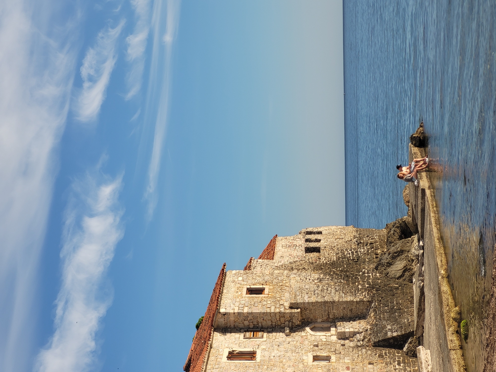 A vibrant, vertically oriented photograph captures a scenic coastal view in Budva, Montenegro, under bright daylight. The left side of the image is dominated by a clear blue sky, adorned with several streaks of wispy white clouds.
On the right, a calm, deep blue sea stretches horizontally across the frame, meeting a faint horizon line. Near the bottom right, two individuals are seen relaxing on a low, paved structure that extends into the water. They appear to be sitting or lounging at the water's edge, partially submerged, enjoying the tranquil setting. One person wears a dark cap.
The lower half and center of the image feature a robust, ancient-looking stone building, likely part of Budva's historic fortifications, with a reddish-brown tiled roof. Constructed from light-colored stone blocks, the building displays several small, rectangular windows. It rises from the rugged, rocky coastline, which forms the base of the structure, and borders the paved area where the people are seated. The overall impression is one of peaceful leisure in a historic Mediterranean environment. No text is visible.