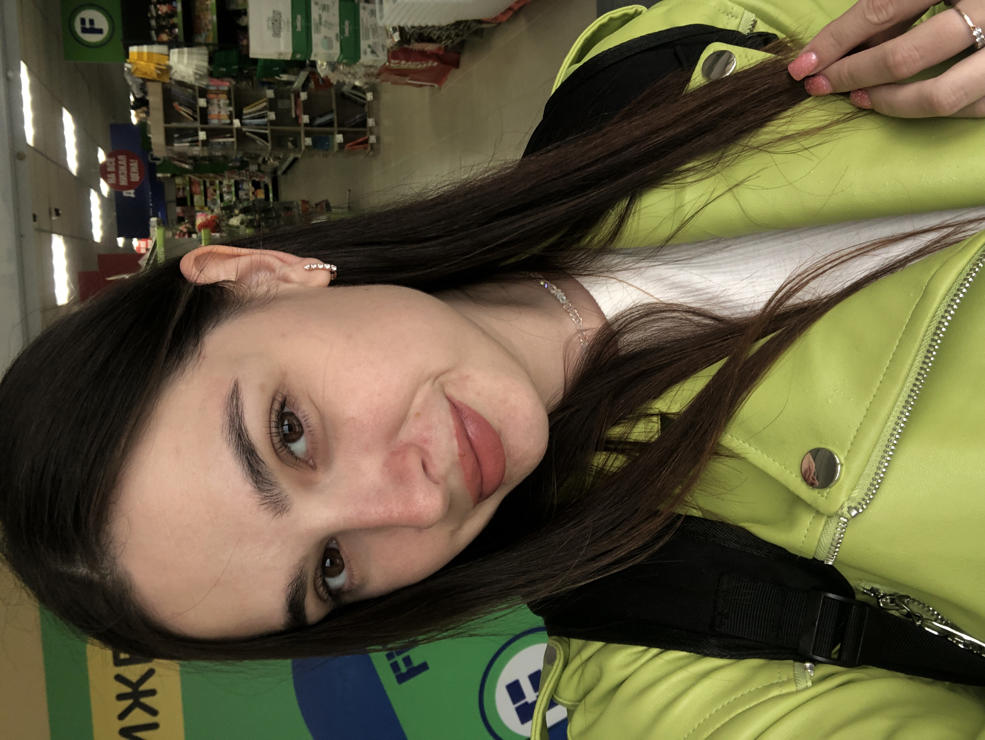 This is a selfie of a young woman in Chaikovsky, Russia. The woman has long, dark brown hair, brown eyes, and light skin. She is wearing a bright green jacket and a white shirt underneath. She has a ring on her left hand with small diamonds. Her nails are painted pink with glitter. A delicate necklace with clear beads is visible around her neck.
The background appears to be an indoor retail environment, possibly a store, with shelves stocked with various items. There are signs with green and blue colors, and some text is visible on these signs, though it is difficult to read clearly. One sign has a green circle with a white "F" inside. Another sign has red lettering within a red circle. The lighting suggests it is daytime. The woman is looking at the camera with a slight smile. The overall mood is casual and personal.