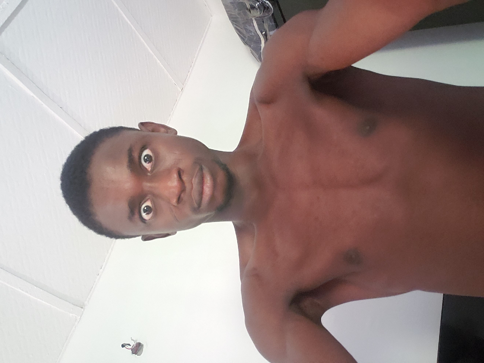 The image captures a young man with dark skin, photographed from a low angle. The picture is rotated 90 degrees counter-clockwise, showing the man's head on the left and his torso extending to the right. He is shirtless, revealing his muscular upper body and arms. His face is turned directly towards the camera, with his eyes wide open, giving an intense, almost startled expression. His lips are slightly parted. He has short, dark hair. The background is primarily a plain white surface, which appears to be a ceiling or an upper wall, with subtle lines indicating paneling or structural elements. In the top right corner of the original image (now the top section of the rotated view), an indistinct dark object, possibly part of a fixture or appliance, is visible. One of the provided crops shows a small opening in the white surface with exposed red and black wires, suggesting an unfinished electrical installation or a simple interior. The lighting is bright and even, consistent with an indoor setting. There is no visible text in the image.