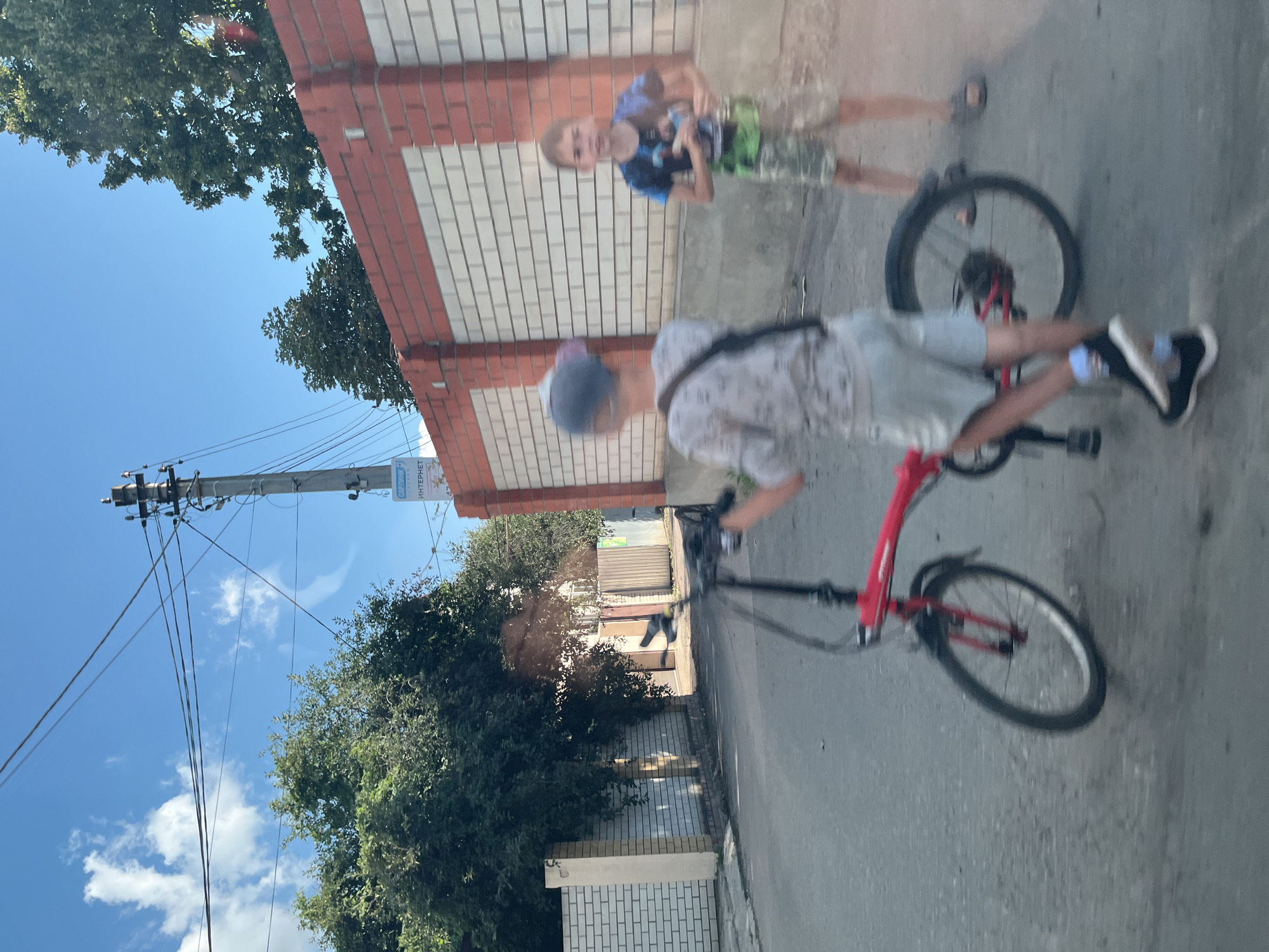 This image depicts two boys on a street in Saratov, Russia. The boy in the foreground, facing away from the camera and slightly to the left, is on a red foldable bicycle. He is wearing a grey t-shirt with a pattern, light-colored shorts, a blue cap, and black sneakers. The boy in the background, facing the camera and slightly to the right, is wearing a blue t-shirt with a graphic design, camouflage shorts, and sandals. He appears to be standing or walking. The street is paved and appears to be a residential area, with brick walls and trees visible on either side. A utility pole with wires is also present. The overall impression is of a casual outdoor scene on a sunny day.
