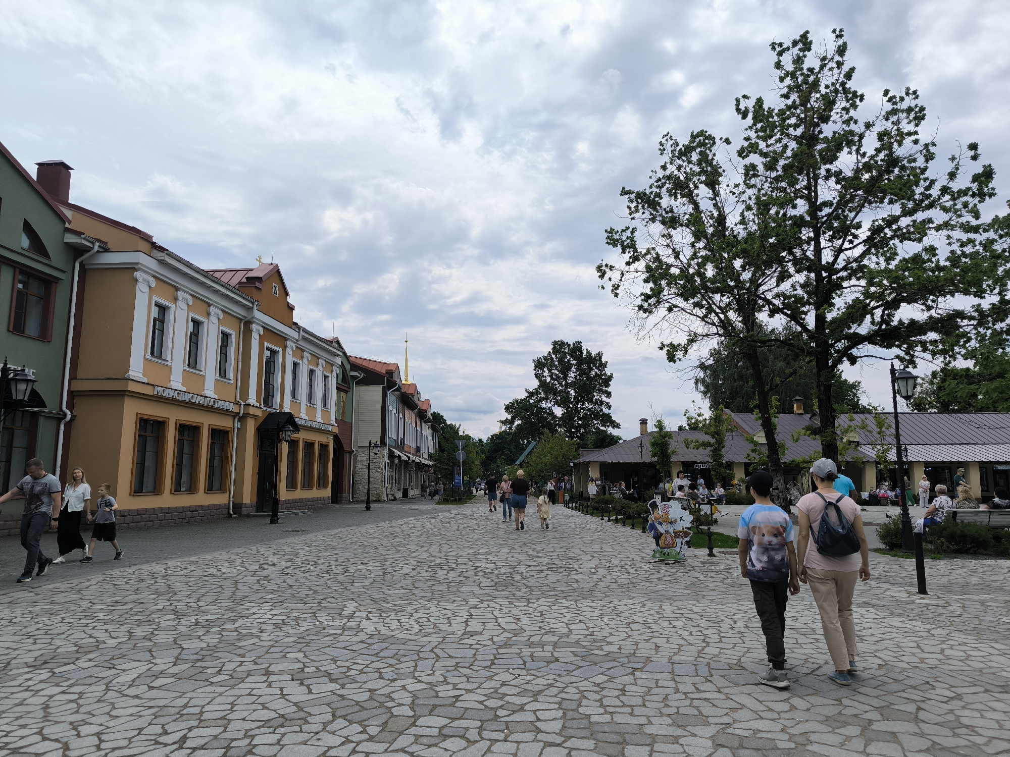 A wide, cobbled pedestrian street lined with colorful buildings is shown under a cloudy sky. Several people are walking along the street, some in small groups and some individually. On the left, a family consisting of a man, a woman, and a young boy are walking away from the camera. Further down the street, more people can be seen walking and some are gathered outside buildings that appear to be shops or restaurants. On the right side of the street, a large tree with green foliage stands prominently. Behind the tree, there are more buildings and outdoor seating areas with people. The architecture of the buildings suggests a European setting, and the general atmosphere is that of a casual daytime outing. The sky is overcast, indicating likely moderate weather, perhaps a bit cool or just before rain. One building on the left has "МОНАСТЫРЬ СВЯТОЙ ГЕОРГИЙ" written on it, which translates to "Monastery of Saint George".